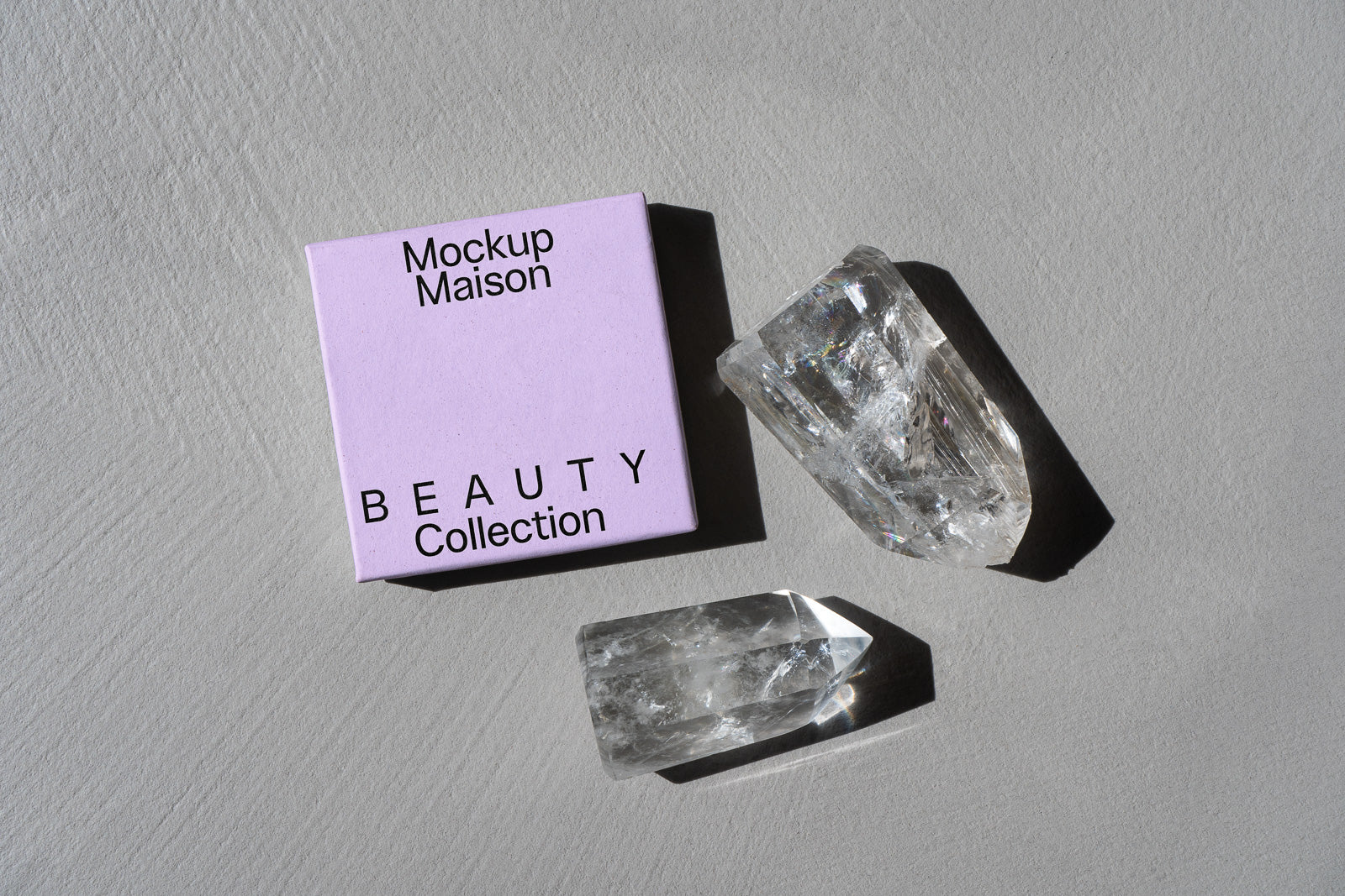 PSD mockup of a light purple beauty collection box labeled "Mockup Maison" next to two clear quartz crystals on a gray textured surface.