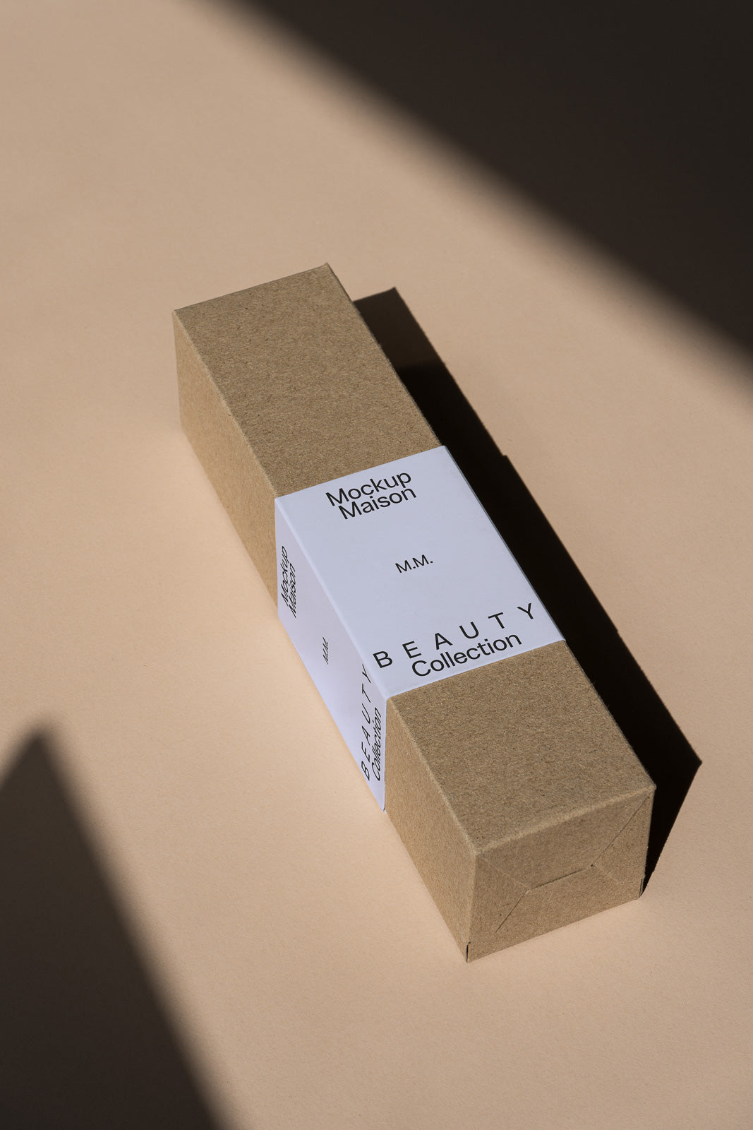 PSD mockup of a rectangular brown cardboard box with a white label titled "Beauty Collection" by Mockup Maison, casting a shadow on a beige surface.