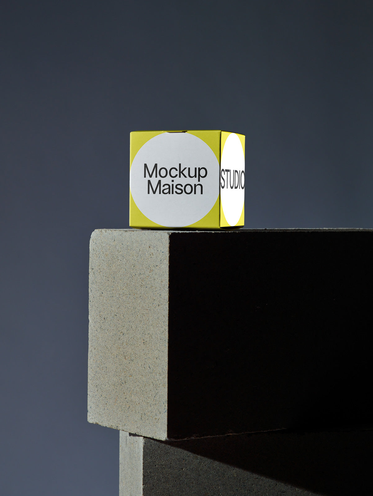 PSD mockup of a yellow cube with circular labels, placed on a concrete block against a dark background, featuring the text "Mockup Maison" and "STUDIO".
