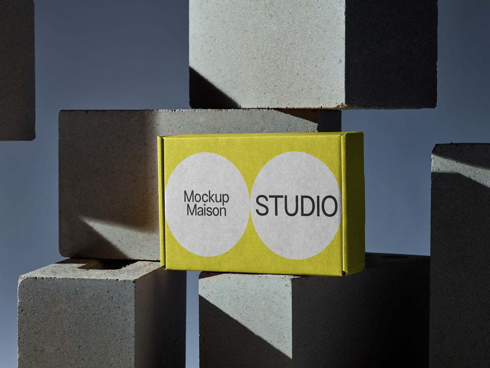 PSD mockup of a yellow rectangular box labeled "Mockup Maison Studio" nestled among large concrete blocks in soft lighting.