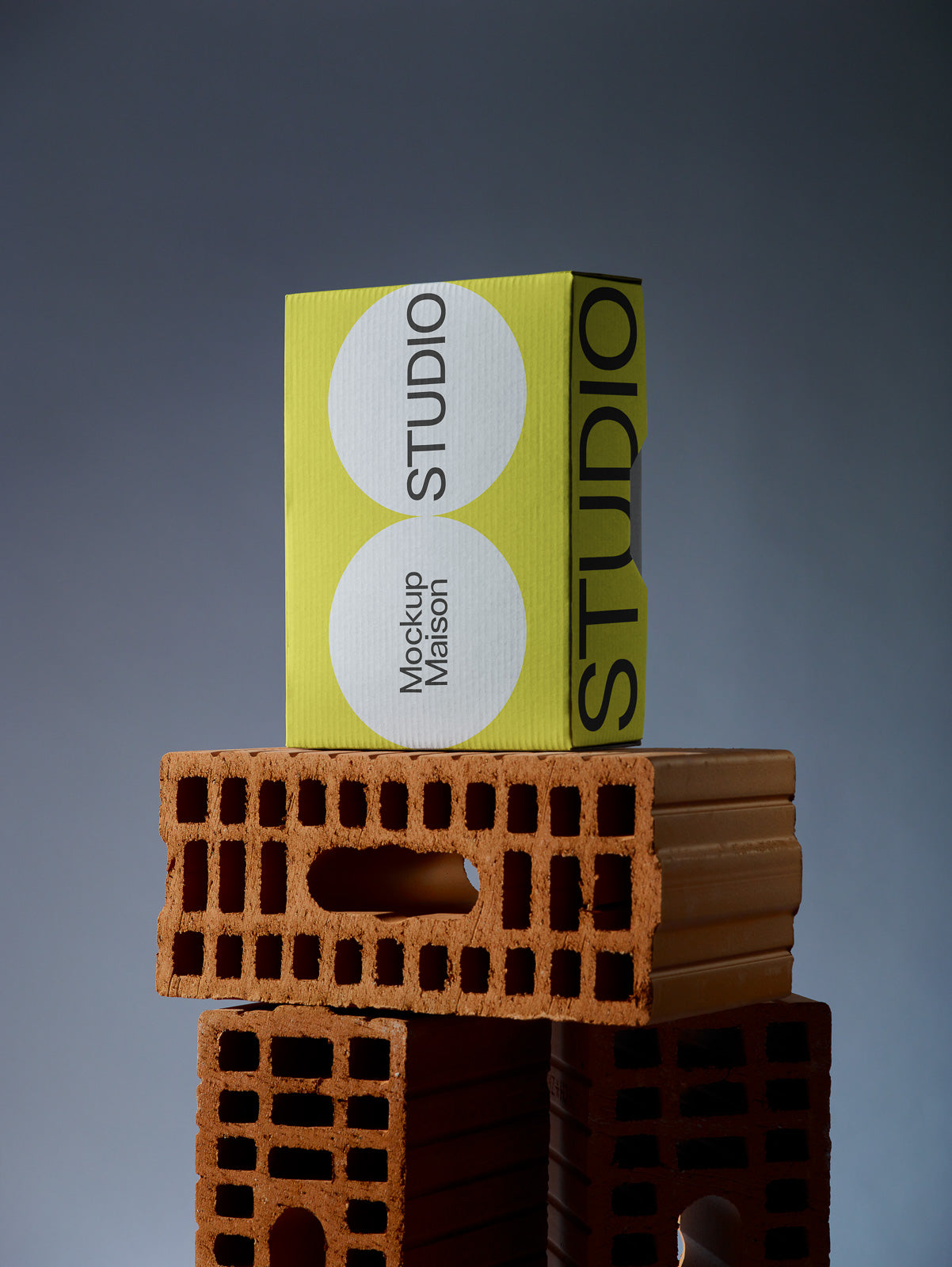 PSD mockup of a yellow rectangular box labeled "STUDIO" and "Mockup Maison," placed on stacked red bricks against a gray background.