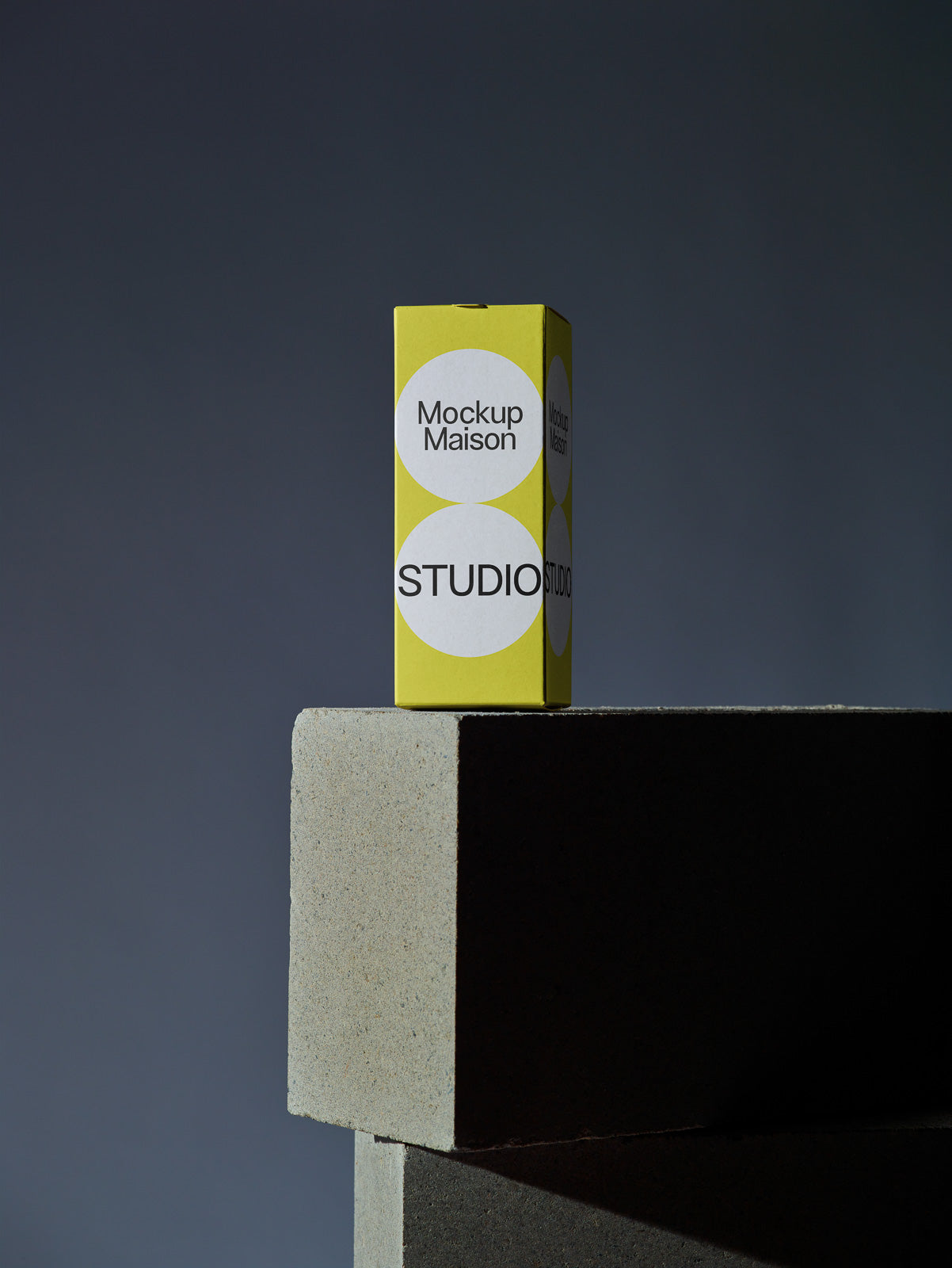 PSD mockup of a yellow rectangular box labeled "Mockup Maison Studio" placed on large geometric concrete blocks against a dark gradient background.