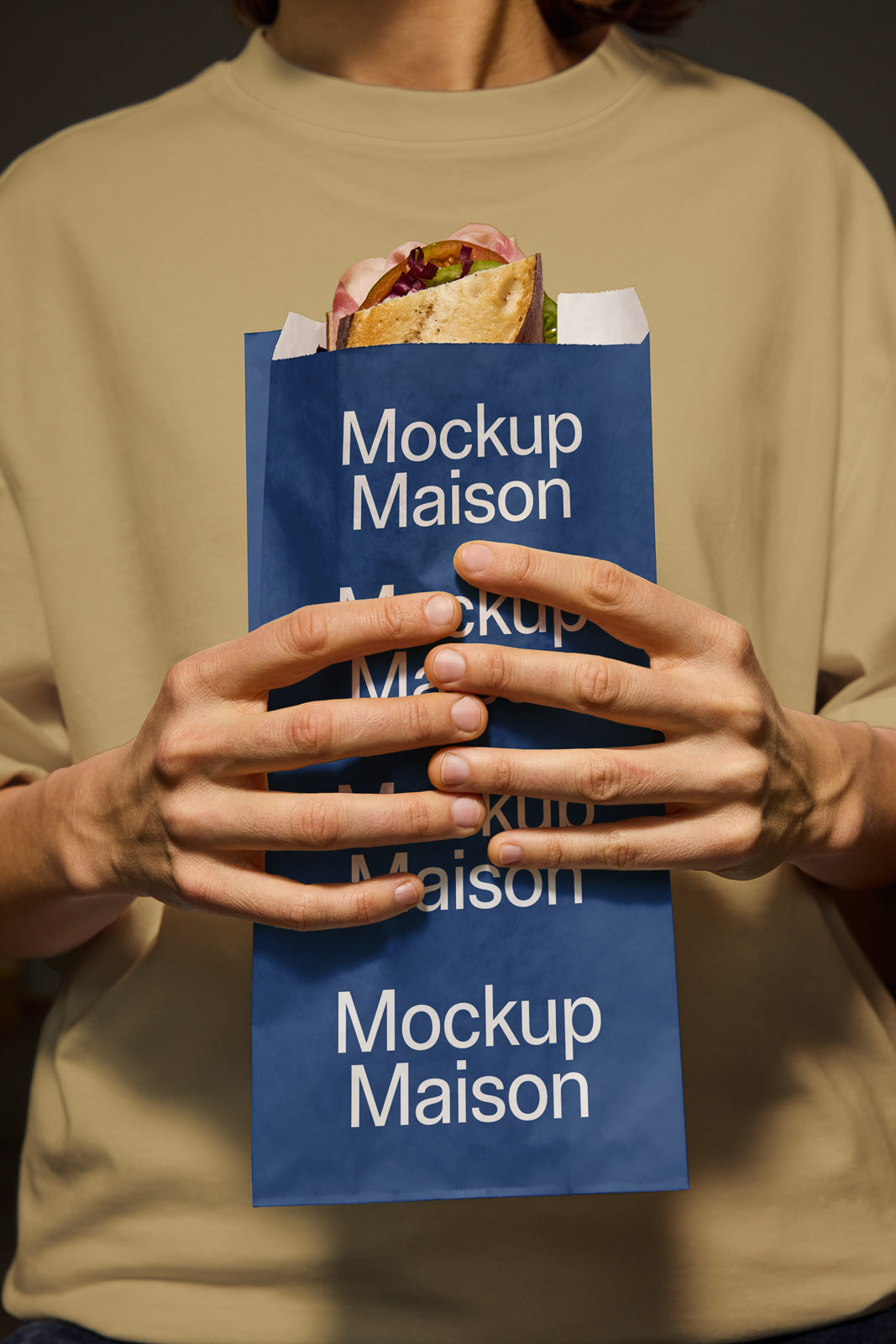 PSD mockup of a person holding a blue paper bag with "Mockup Maison" text, containing a sandwich, against a beige shirt background.