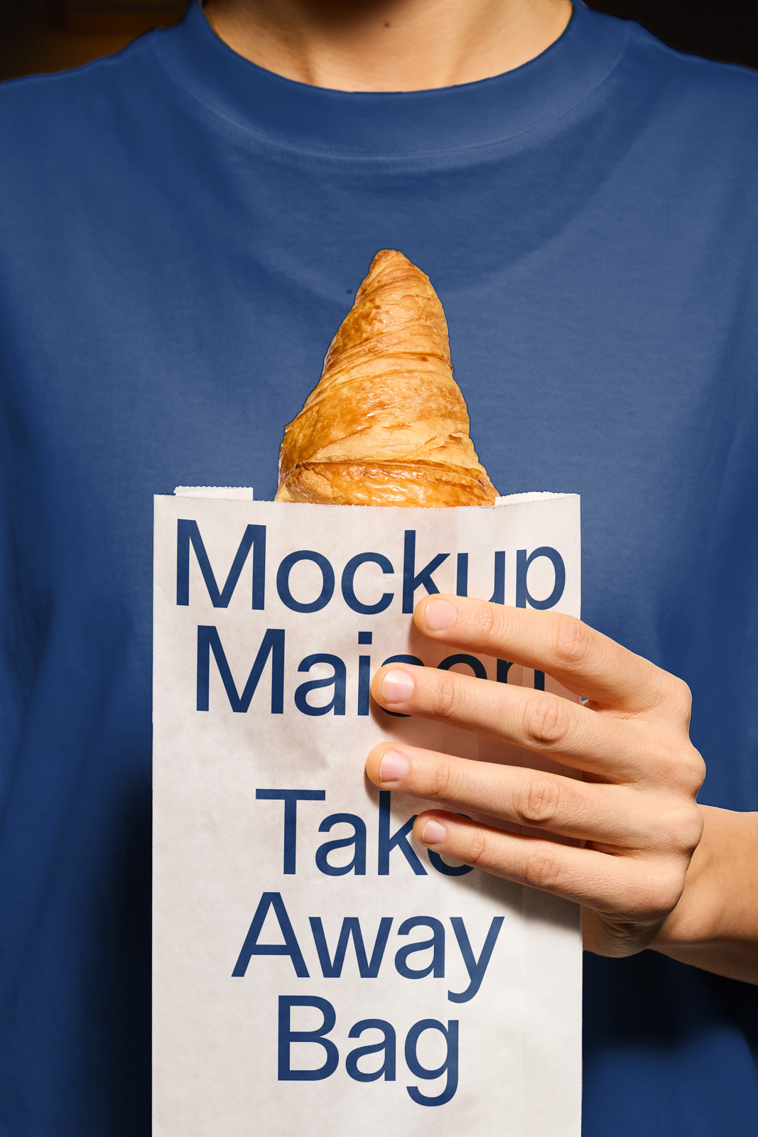 PSD mockup of a hand holding a paper bag labeled "Mockup Maison Take Away Bag," with a croissant partially visible from the top.