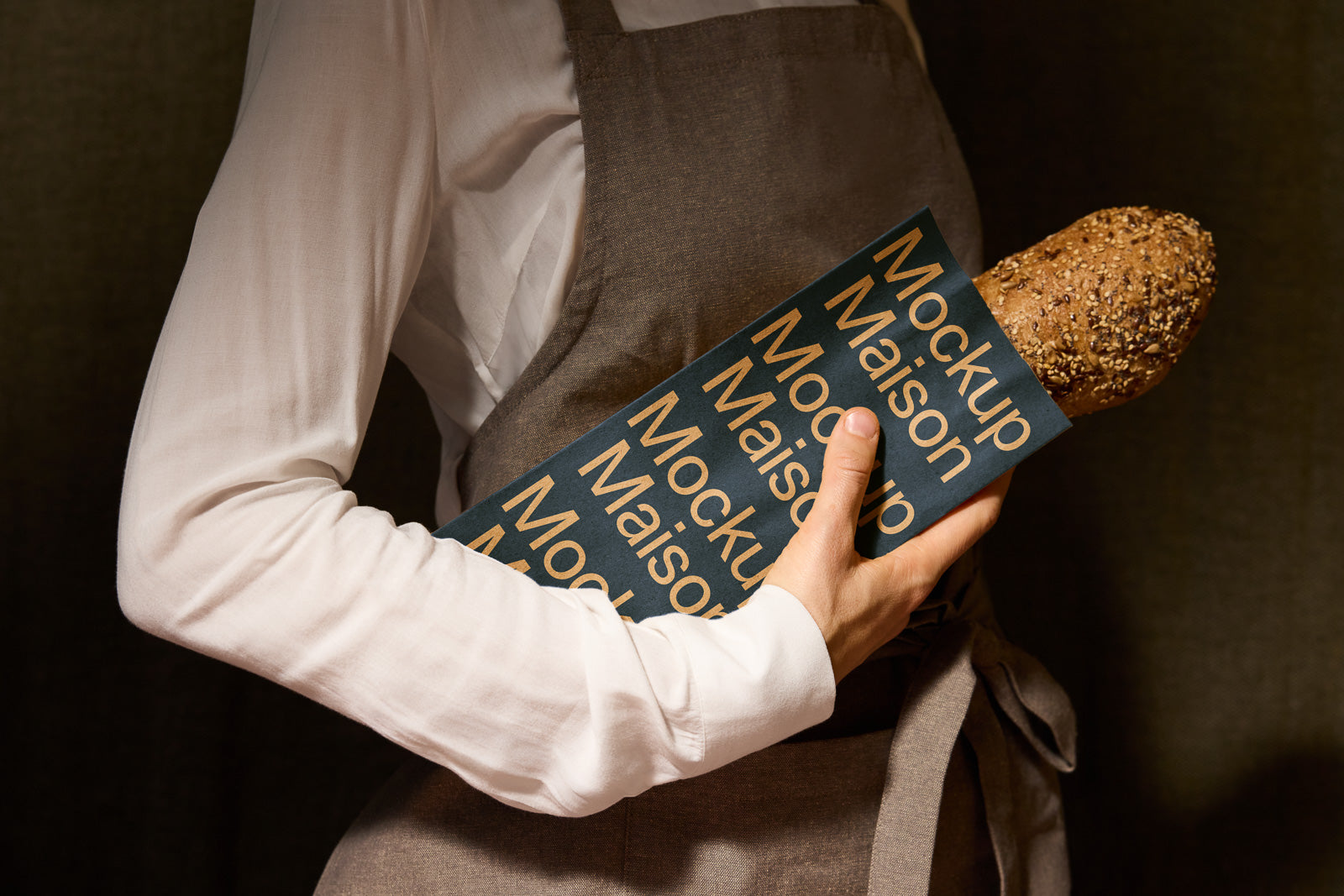PSD mockup of a person in a white shirt and apron holding a baguette in a blue paper sleeve labeled "Mockup Maison" against a dark background.