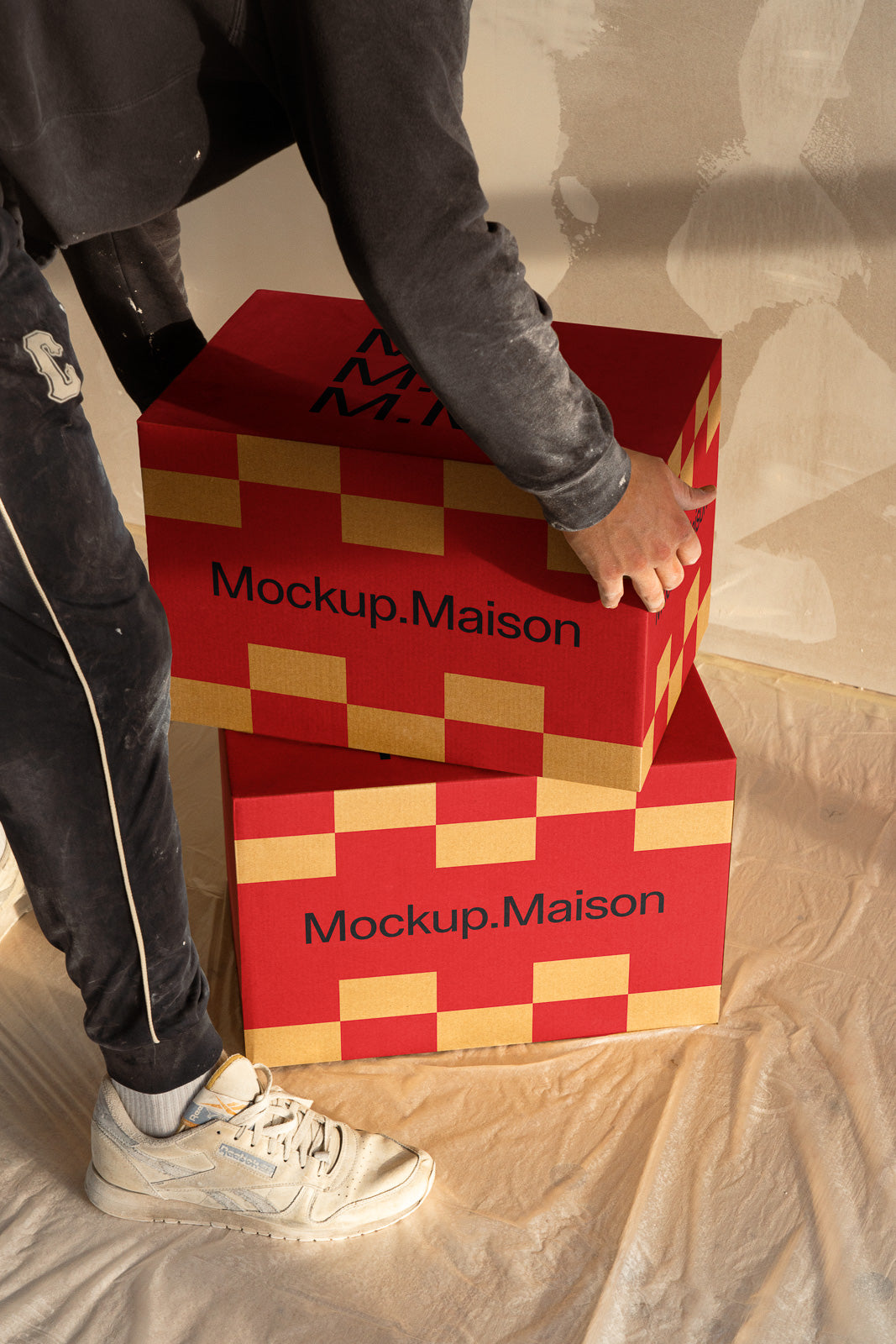 PSD mockup of a person stacking two red and yellow cardboard boxes labeled "Mockup.Maison" on a covered floor, with detailed ambient lighting.