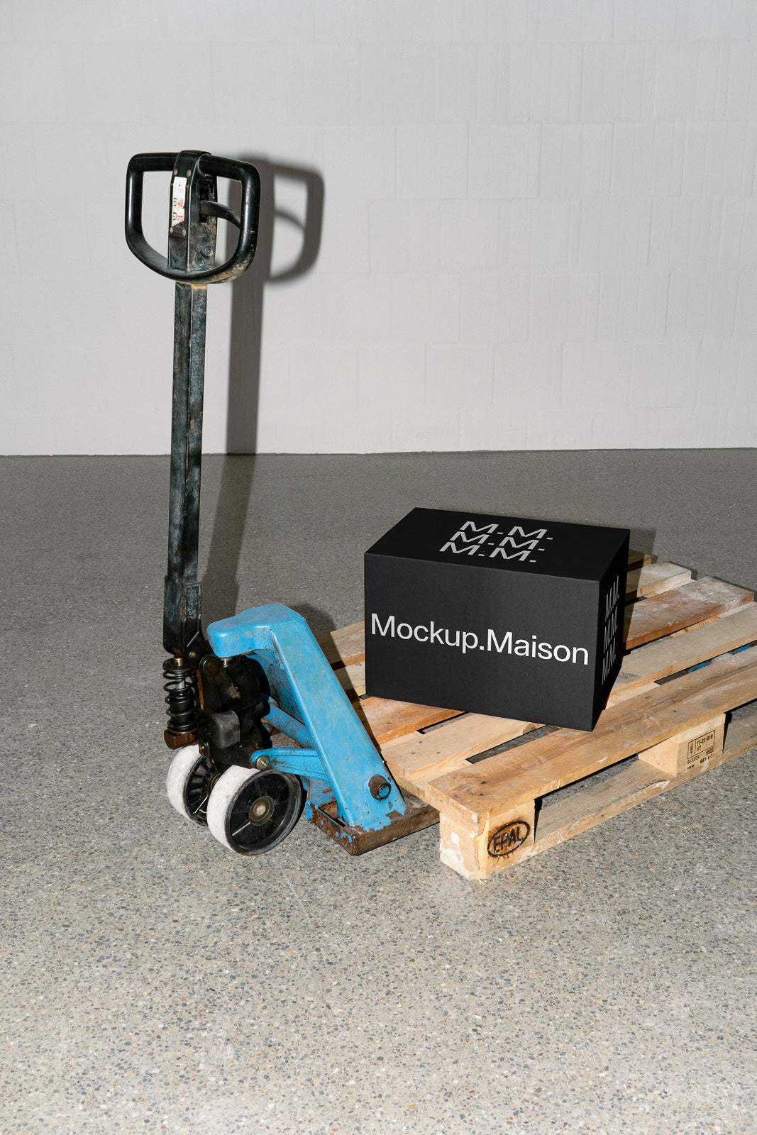 PSD mockup of a black box labeled "Mockup.Maison" placed on a wooden pallet beside a blue manual pallet jack in an industrial setting.