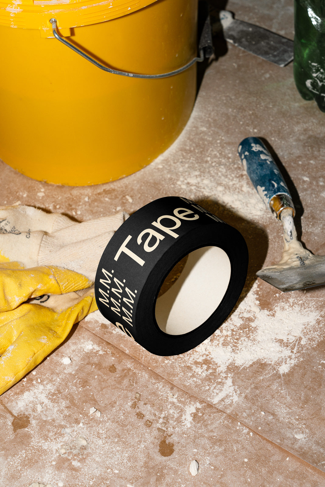 PSD mockup of black tape roll with white text lying on dust-covered floor, near yellow bucket, paint scraper, and gloves.