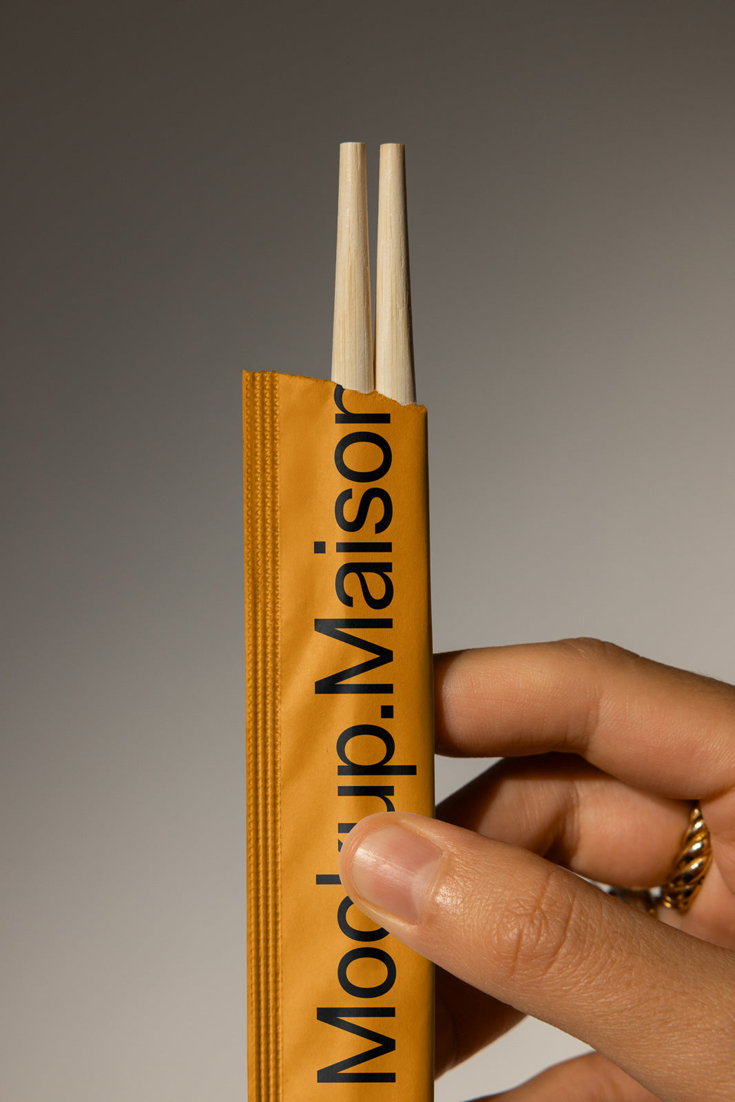 PSD mockup of a hand holding a yellow paper sleeve with wooden chopsticks partially exposed, featuring bold black typography on the sleeve.