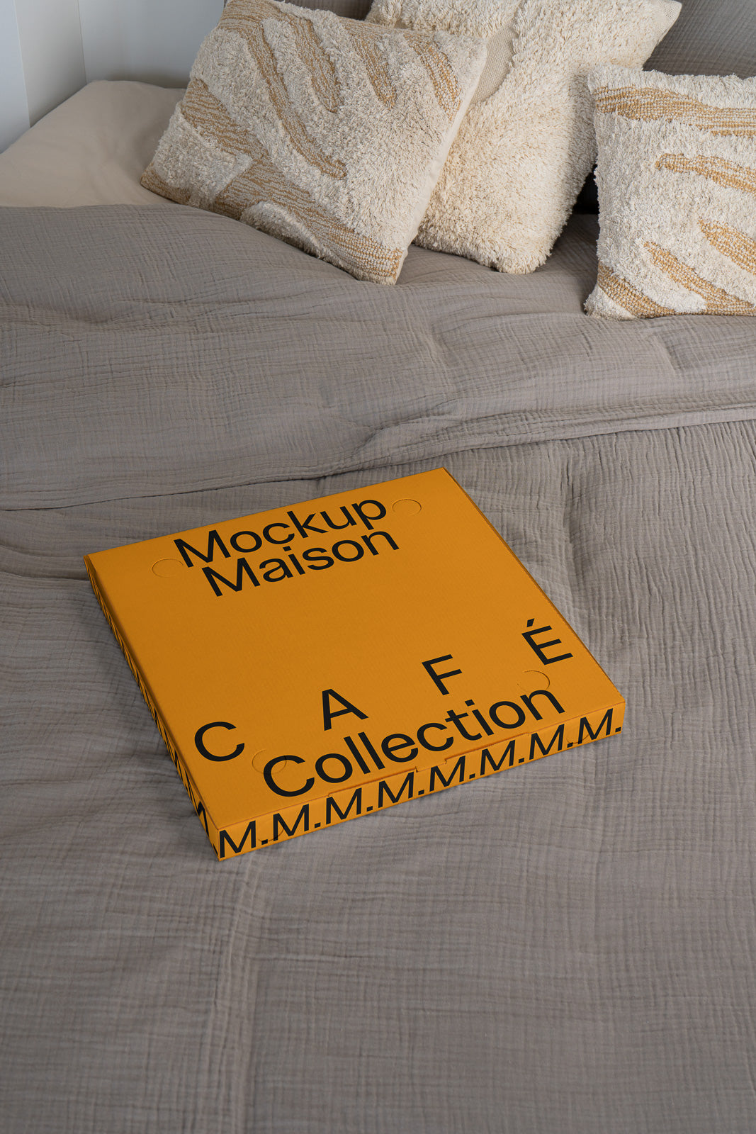 PSD mockup of a square orange packaging box labeled "Mockup Maison Café Collection" resting on a textured gray bedspread with patterned pillows.