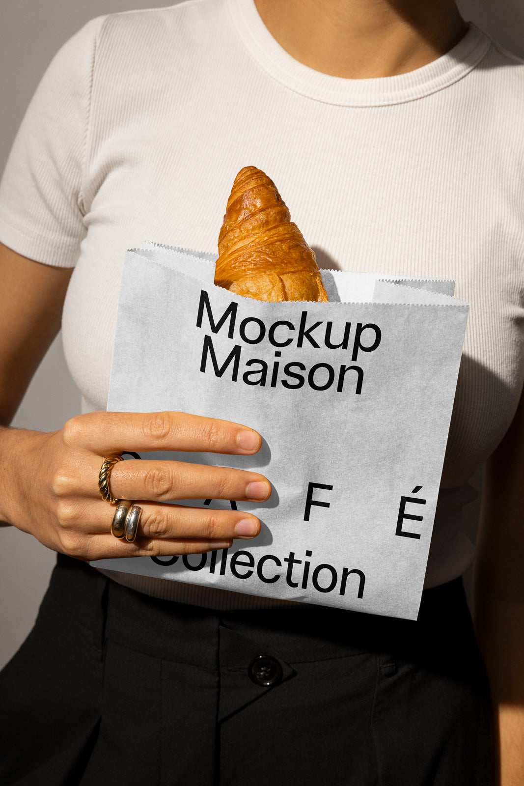 PSD mockup of a woman holding a paper bag labeled "Mockup Maison Café Collection" with a croissant peeking out, wearing a white shirt and rings.