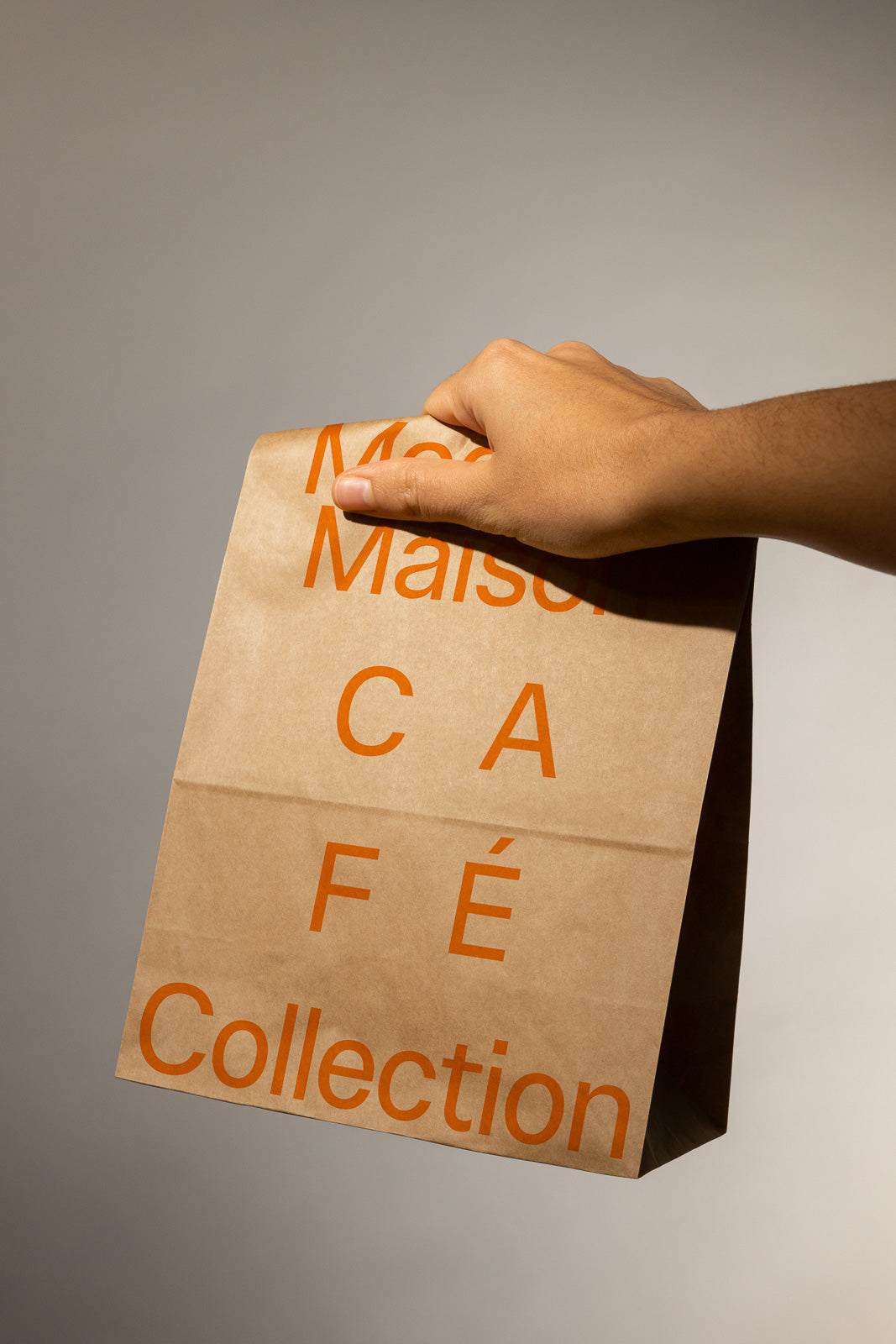 PSD mockup of a hand holding a brown paper bag with orange text that reads "Maison Café Collection" on a neutral background.