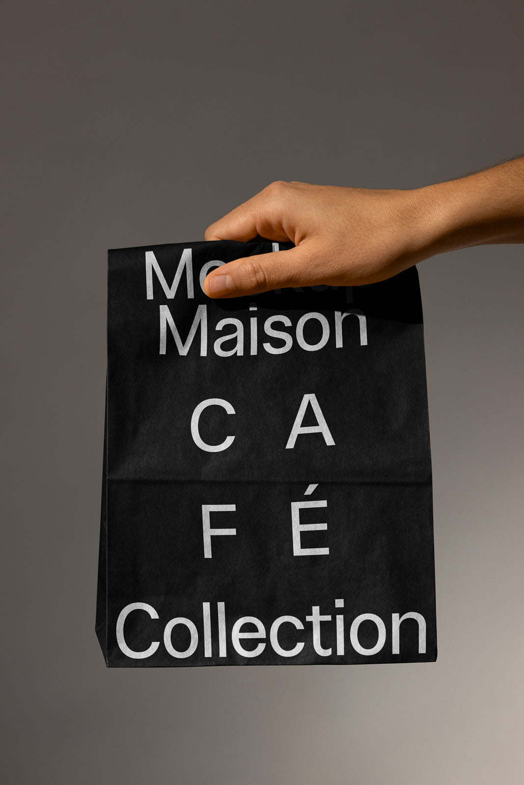 PSD mockup of a hand holding a black paper bag with white text reading "Maison Café Collection."