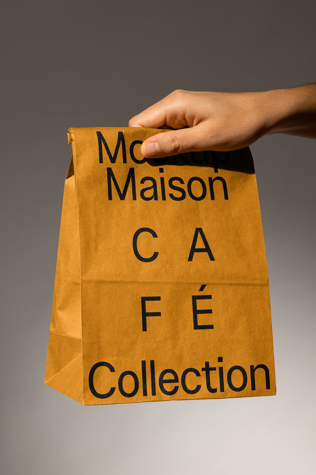 PSD mockup of a hand holding a brown paper bag labeled "Mockup Maison Café Collection" with bold black text, set against a neutral background.