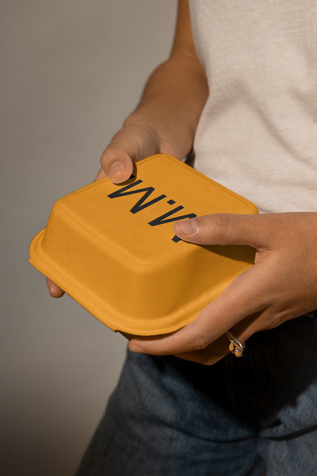 PSD mockup of a person holding a closed yellow textured food container with bold black text, wearing a white shirt and jeans.