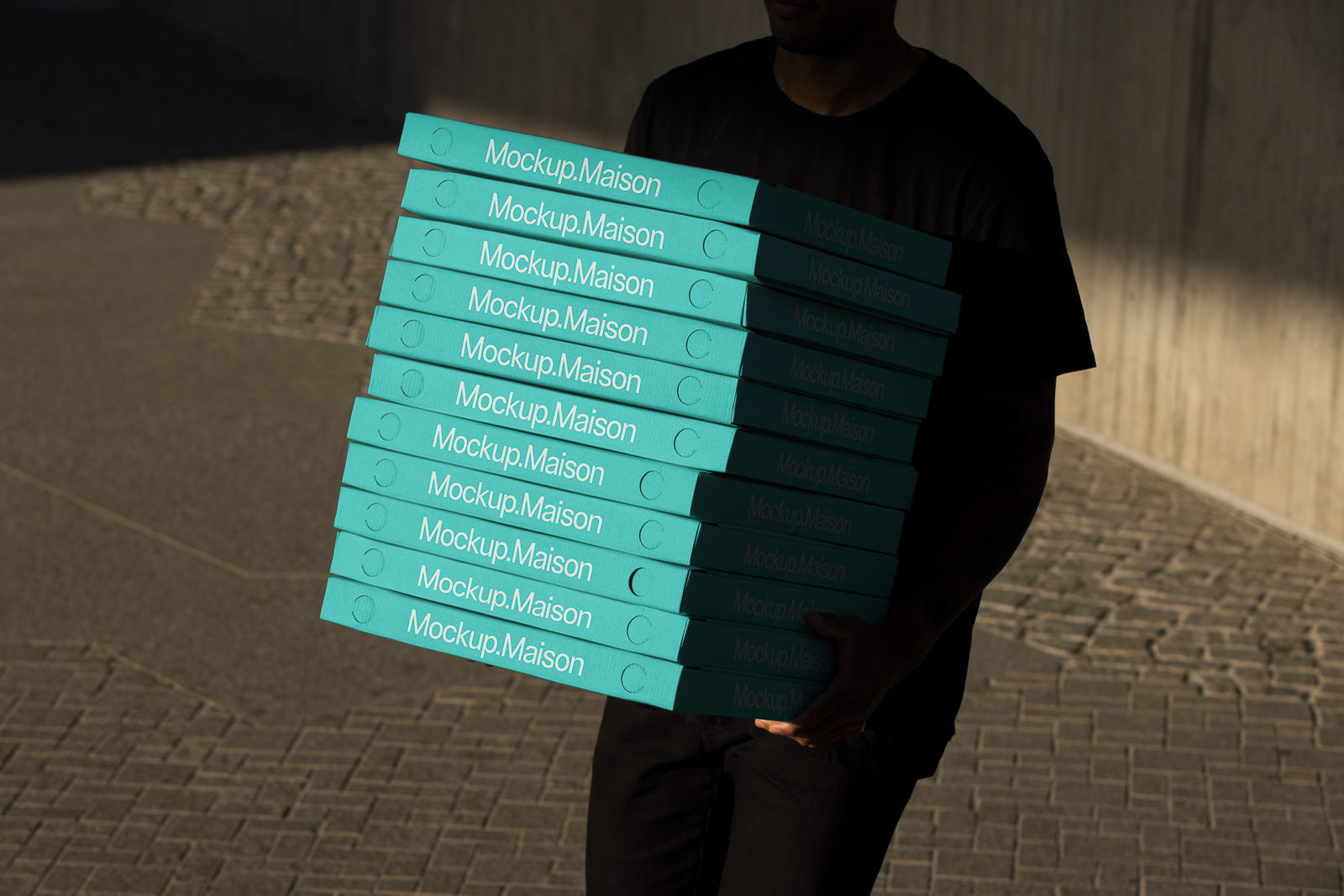 PSD mockup of a person carrying a stack of teal boxes labeled "Mockup.Maison" in an outdoor setting, with cobblestone paving in the background.
