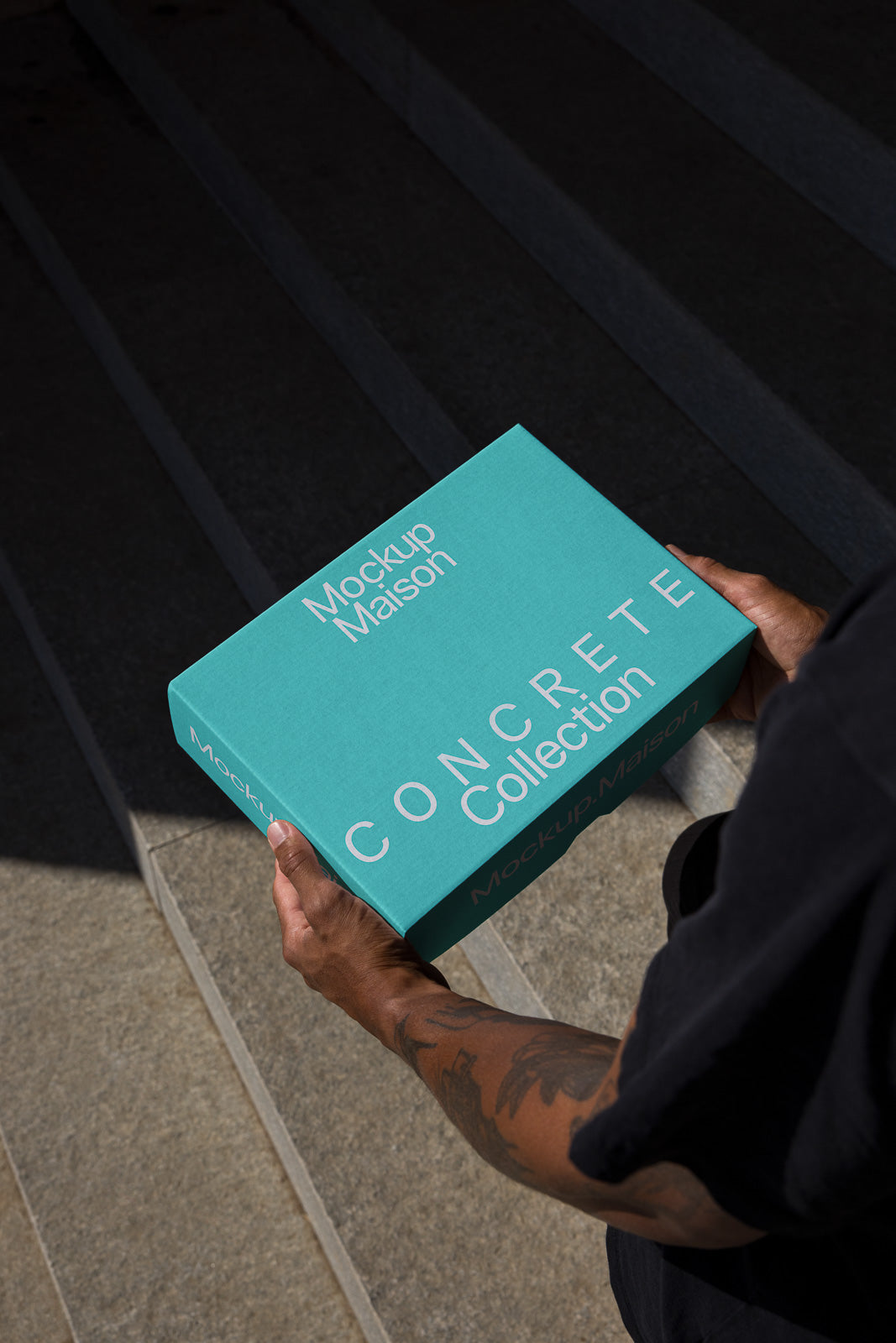 PSD mockup of hands holding a turquoise box labeled "Mockup Maison CONCRETE Collection" outdoors on a stone stairway.