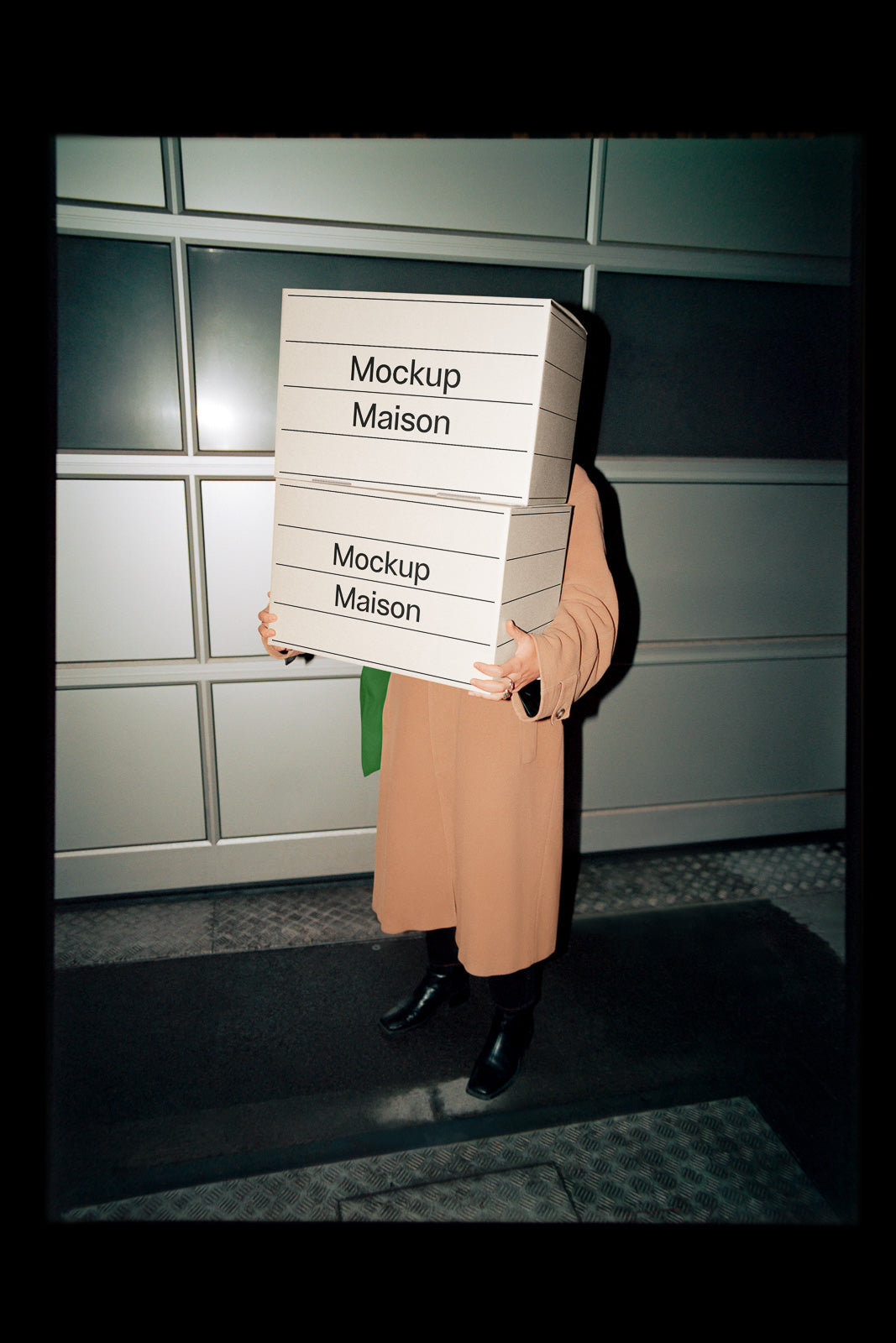 PSD mockup of a person holding two stacked boxes labeled "Mockup Maison," standing against a modern, tiled wall while wearing a long beige coat.