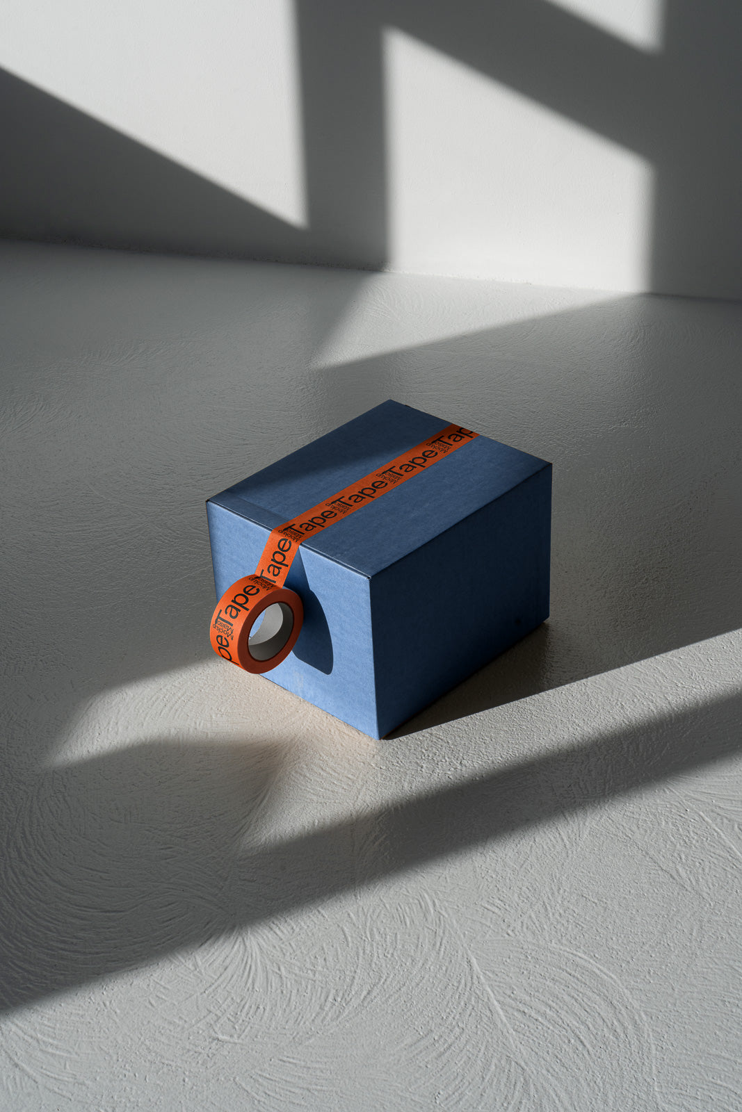 PSD mockup of a blue textured box with red tape partially wrapped around it, casting distinct geometric shadows on a light surface.
