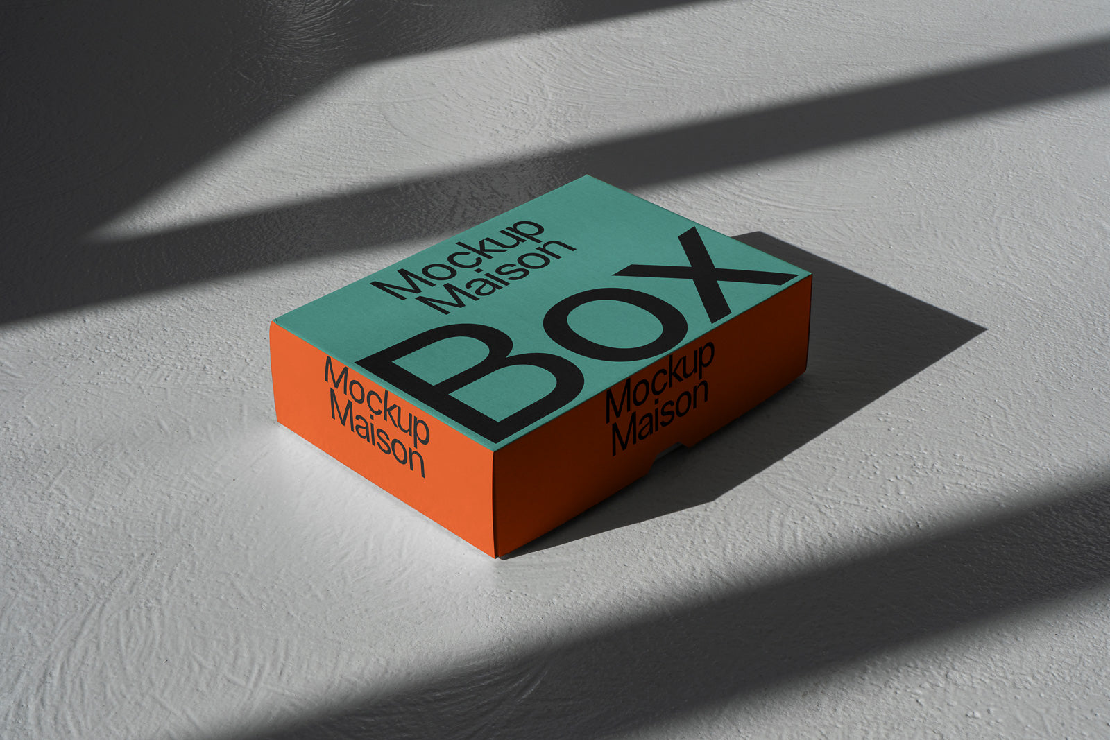 PSD mockup of a box with teal top and orange sides, displaying the text "Mockup Maison" and "BOX" in bold black letters, casting a shadow on a textured surface.