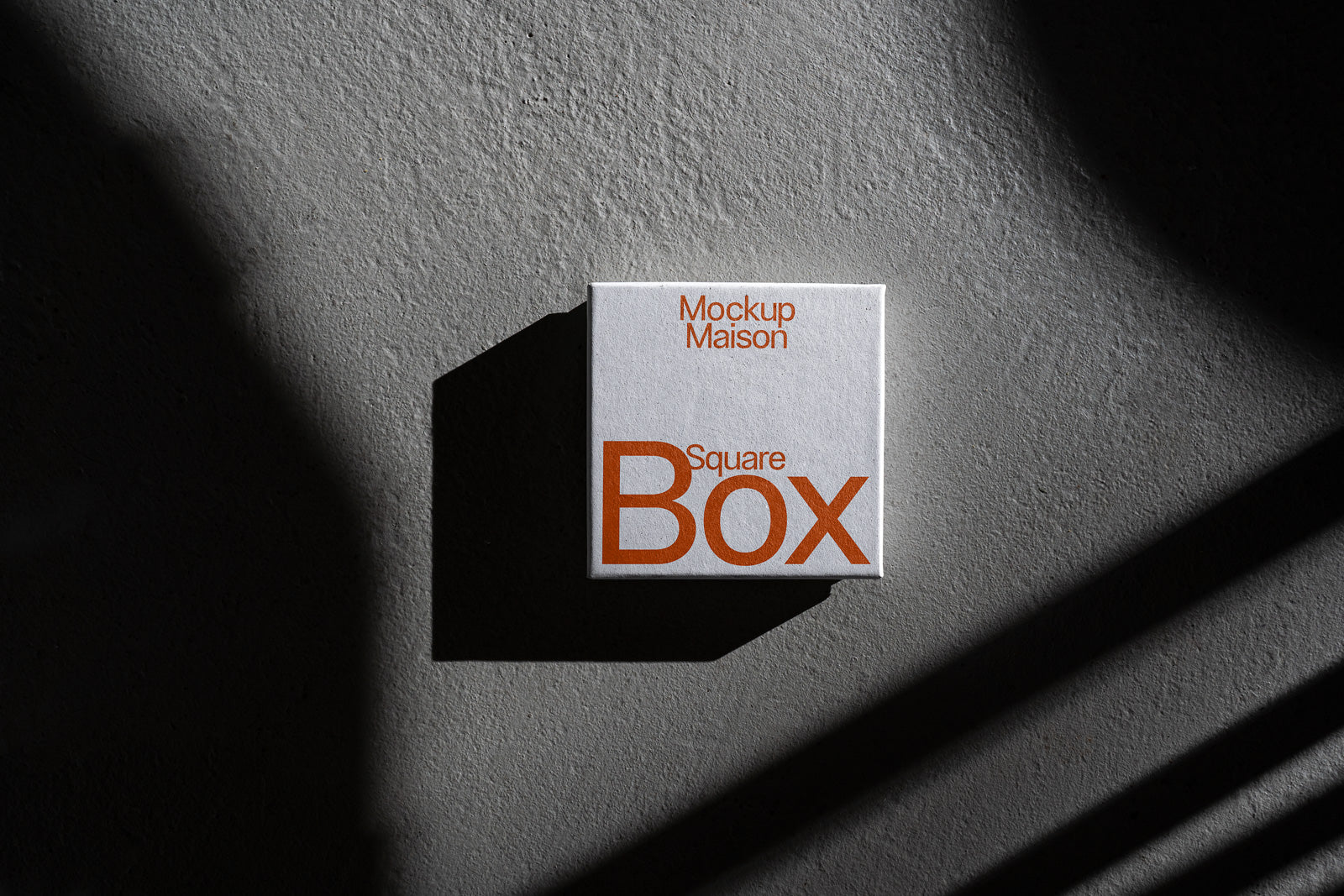 PSD mockup of a square box with the text "Mockup Maison Square Box" casting a shadow on a textured gray surface.