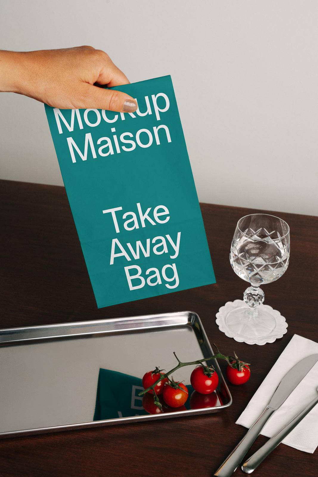 PSD mockup of a teal take-away bag labeled "Mockup Maison" held by a hand, next to a tray with cherry tomatoes and a glass on a table.
