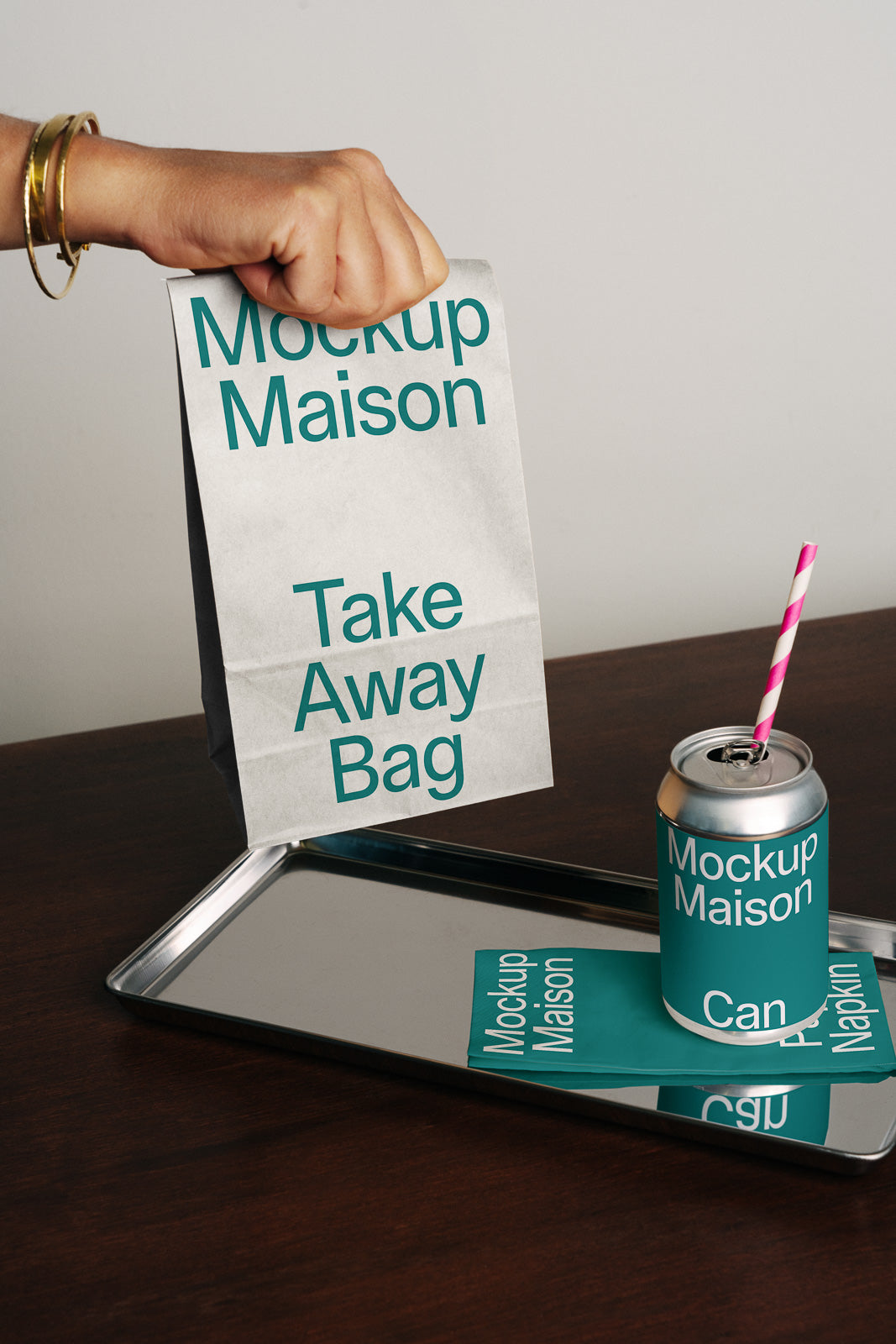 PSD mockup of a hand holding a takeaway bag labeled "Mockup Maison" next to a can with a straw and matching napkins on a metal tray.