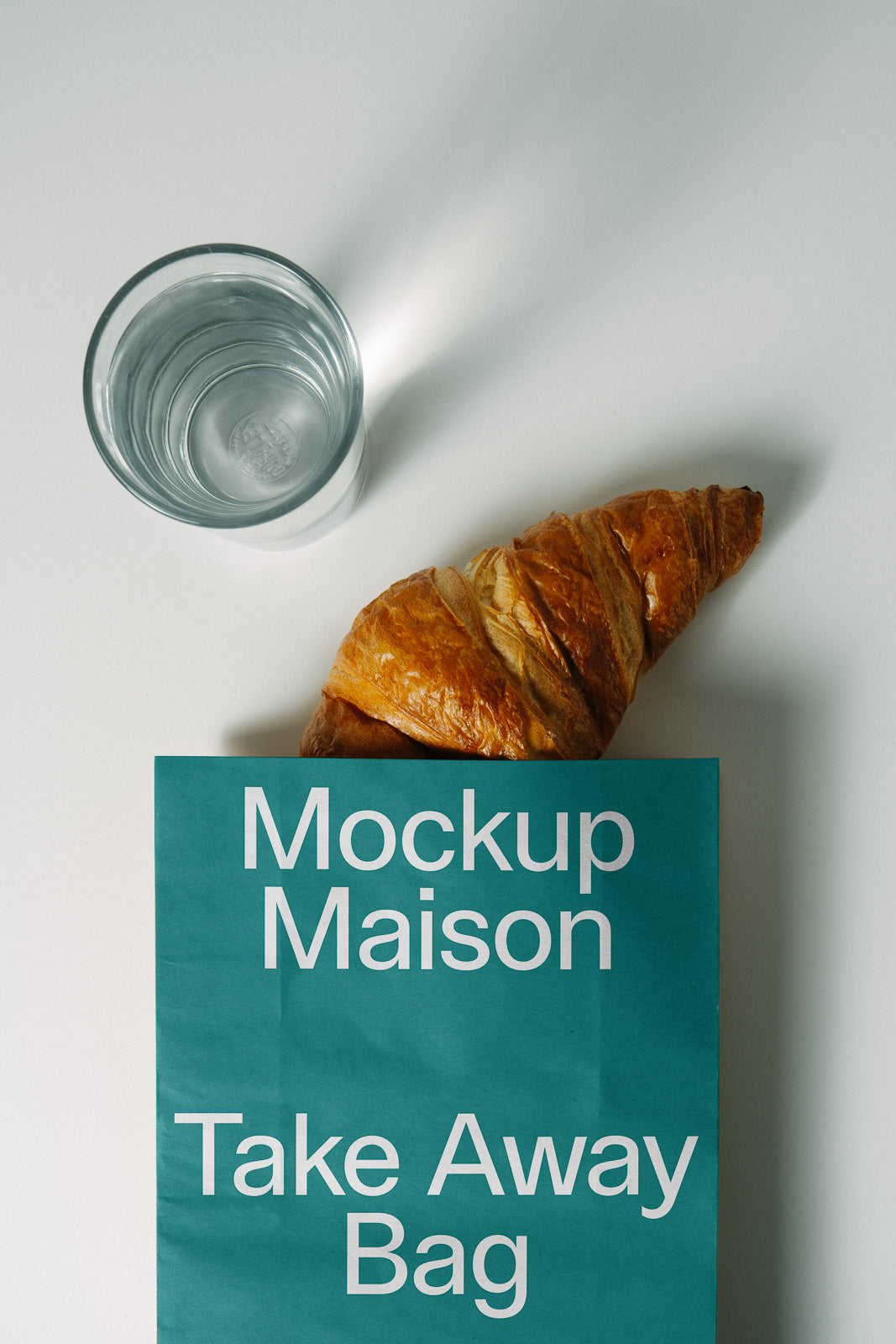 PSD mockup of a "Mockup Maison Take Away Bag" on a table, beside a glass of water and a croissant.