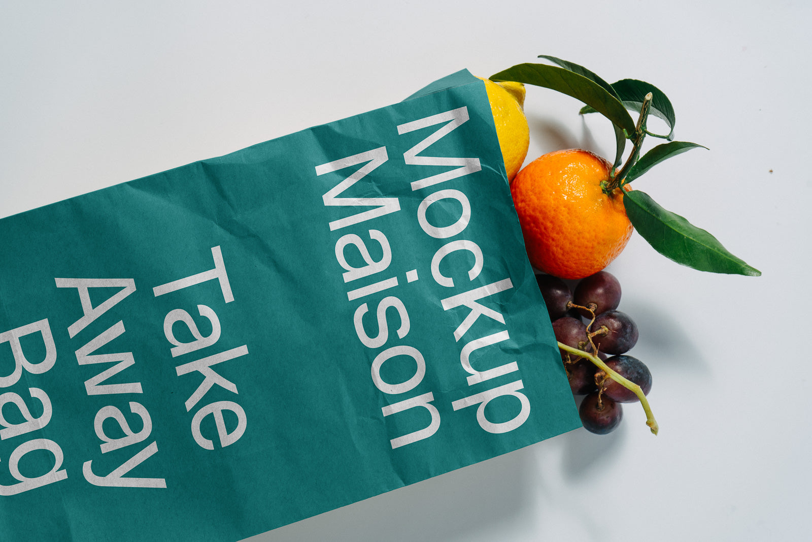 PSD mockup of a teal paper bag with “Mockup Maison” text partially filled with fresh fruits, including lemon, orange, and grapes.