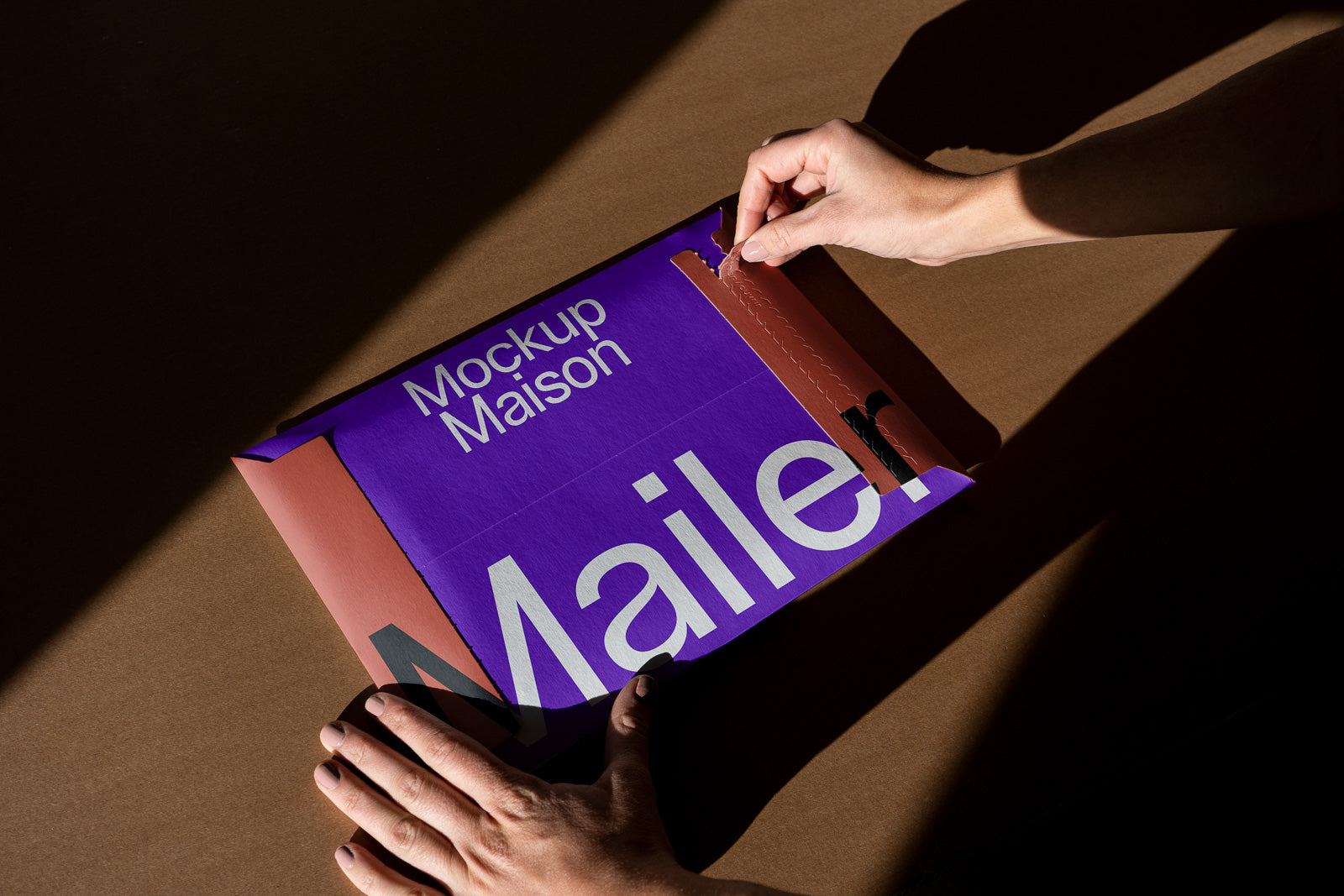PSD mockup of a person opening a purple and brown mailer envelope labeled "Mockup Maison Mailer" on a dark surface.