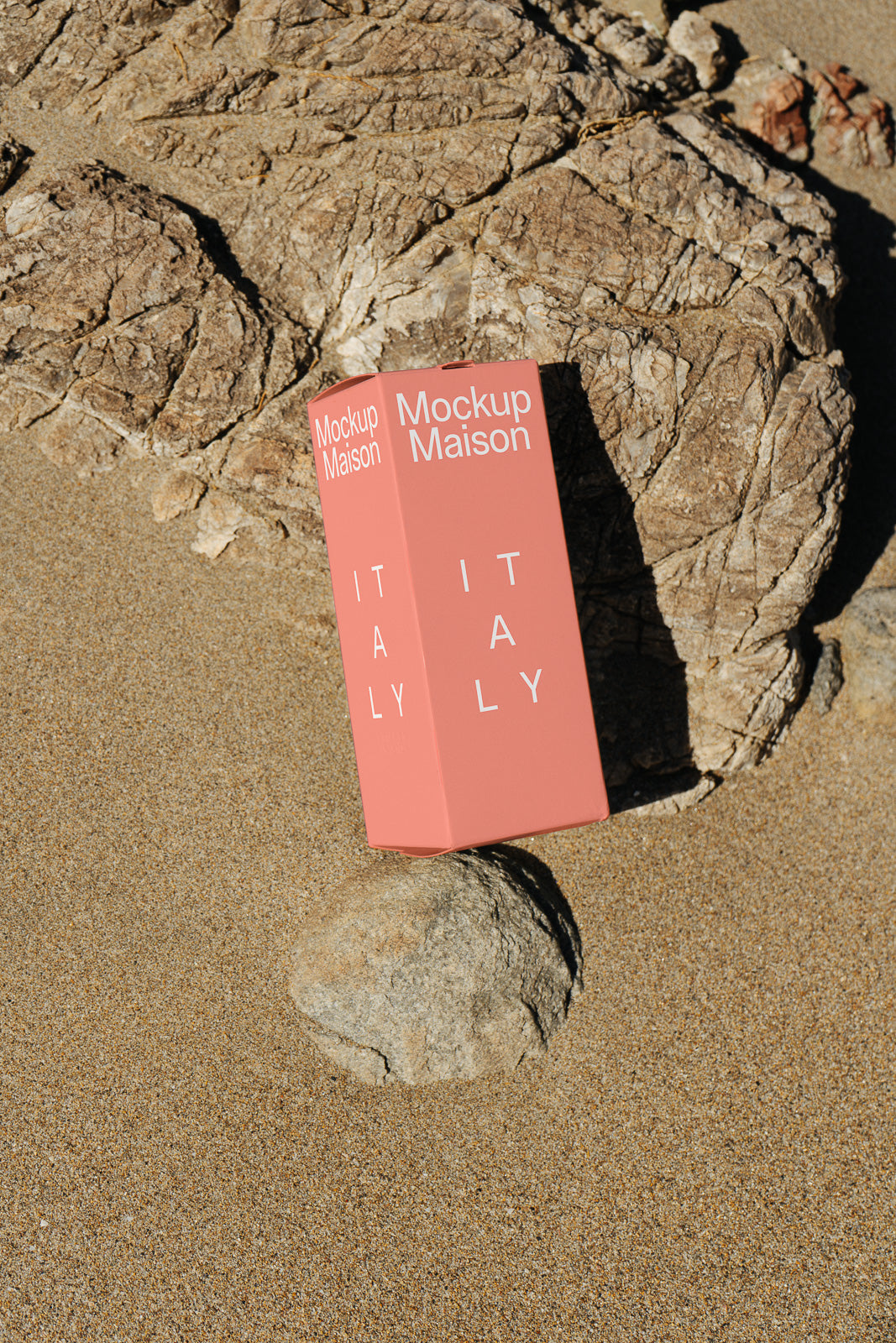 PSD mockup of a coral-colored packaging box labeled "Mockup Maison" and "Italy," placed on sandy terrain beside a large rock.