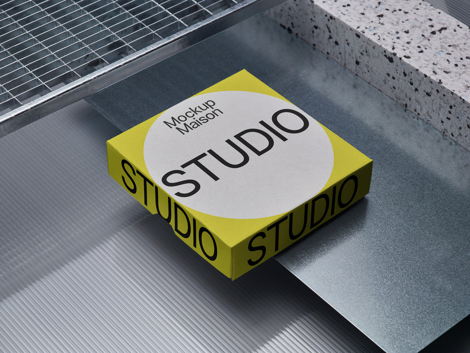 PSD mockup of a yellow box labeled "STUDIO" with a circular white label, placed on a metallic surface surrounded by textured materials.