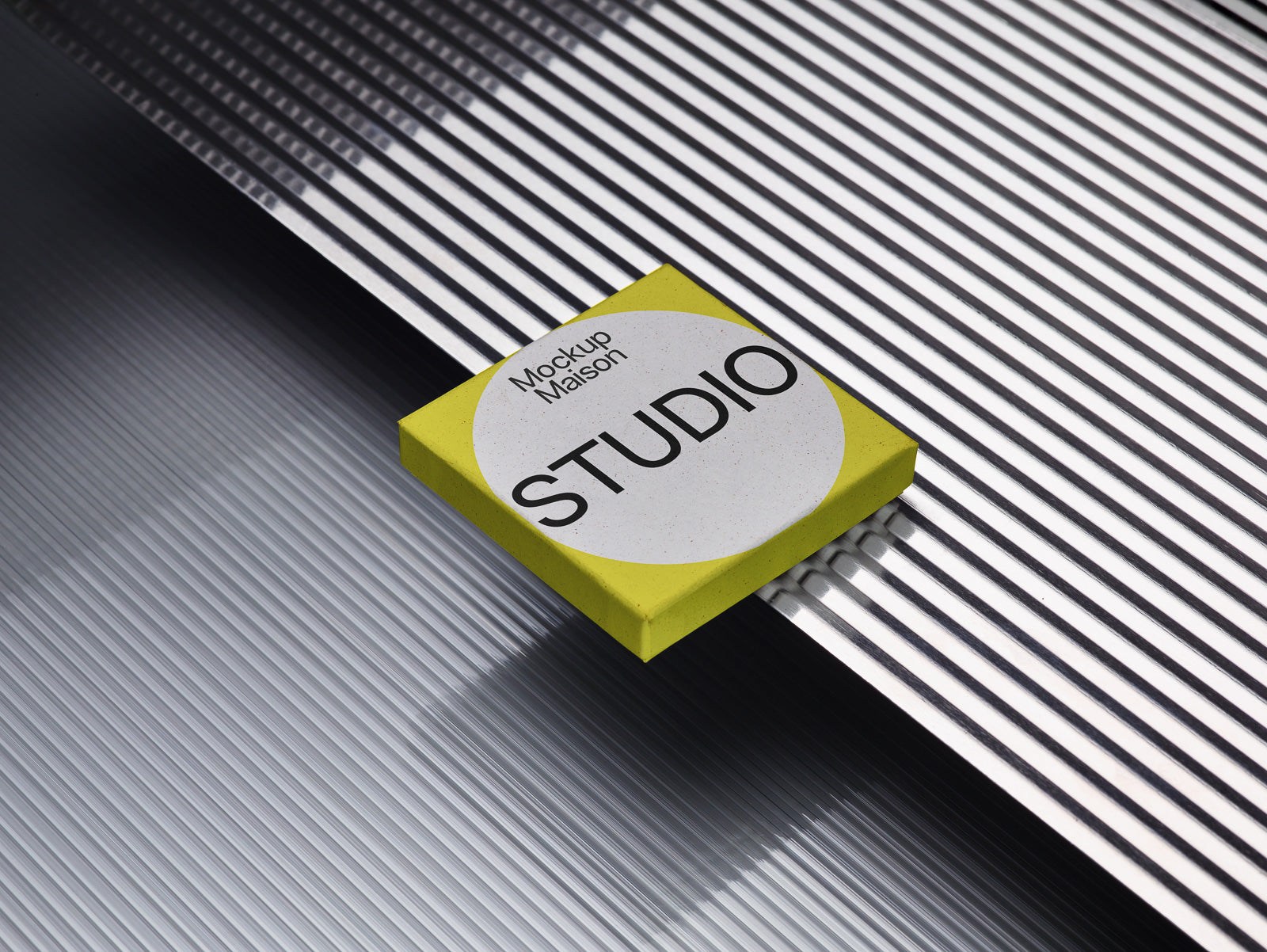 PSD mockup of a square, yellow box with the text "Mockup Maison STUDIO" on a textured, ridged metal surface.