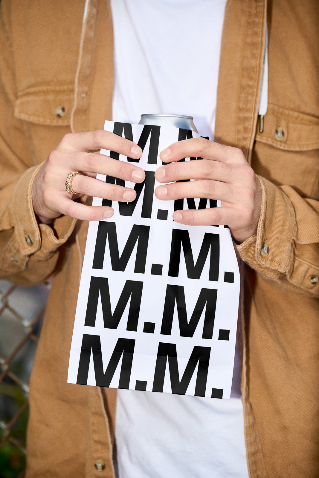 PSD mockup of hands holding a paper-wrapped can, showcasing repetitive black "M.M." text design on a white background.