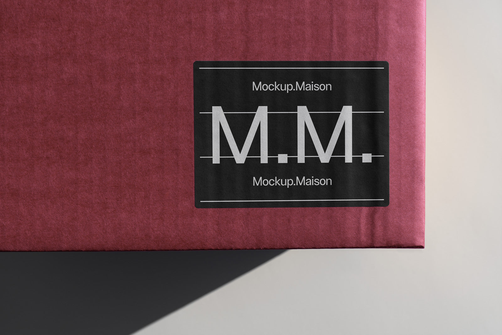 PSD mockup of a burgundy textured box featuring a prominent black and white label with the letters "M.M." and the text "Mockup.Maison" printed on it.