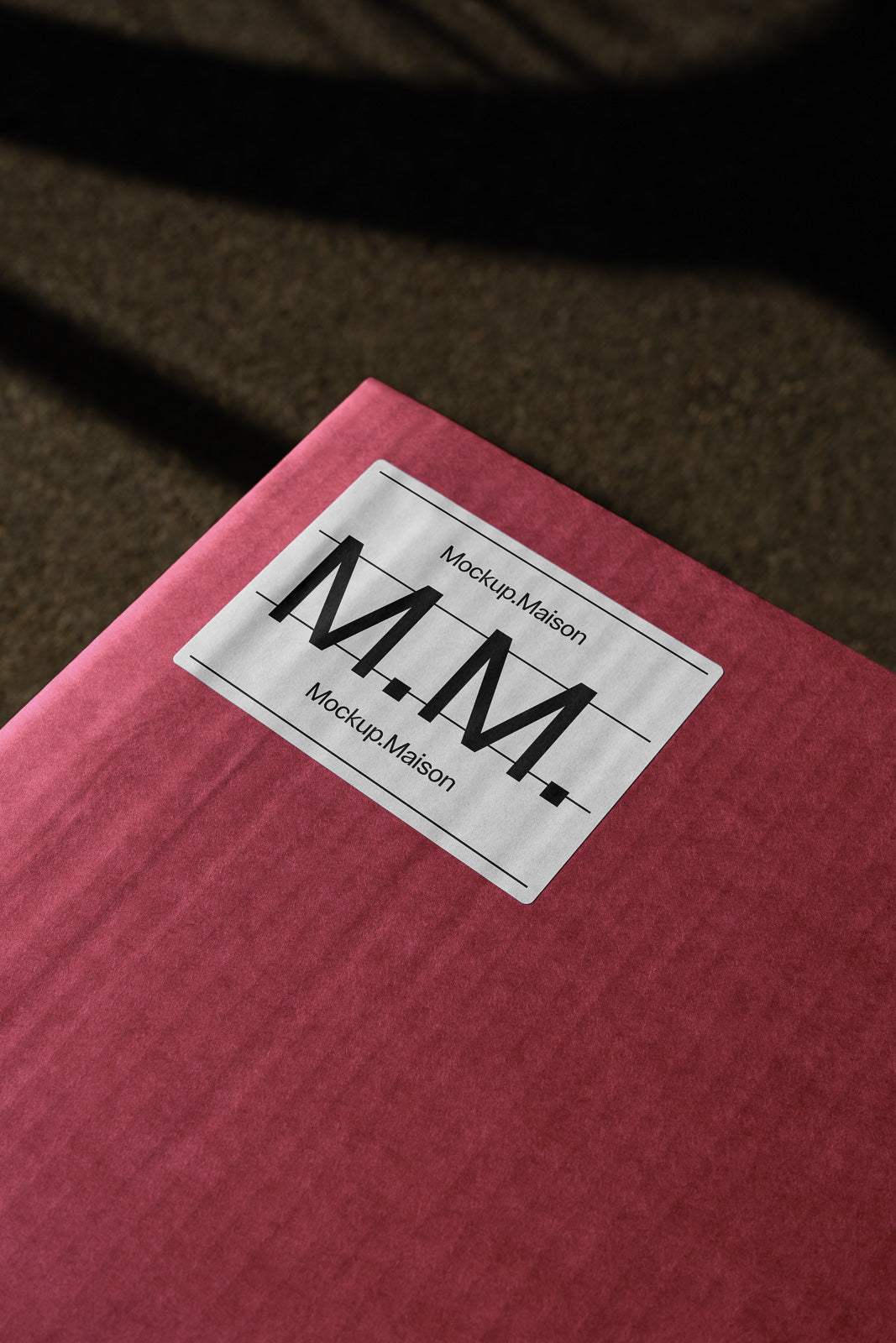 PSD mockup of a red package with a white label displaying the text "Mockup Maison" and "M.M.", placed on a textured surface with shadows.