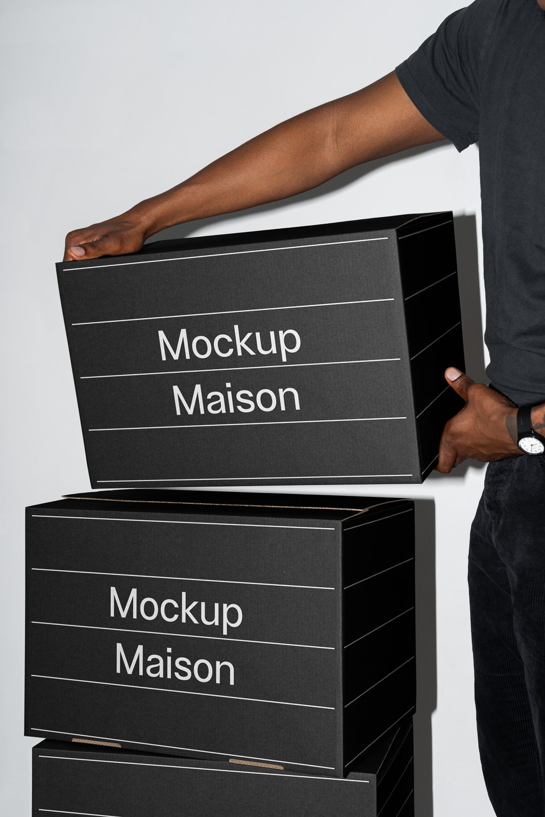 PSD mockup of black cardboard boxes labeled 'Mockup Maison' being held by a person in a black shirt, showcasing a versatile packaging design.