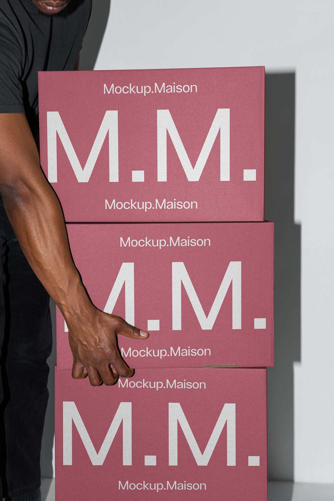 PSD mockup of stacked pink boxes with large white "M.M." text, partially held by a hand, branded with "Mockup.Maison".