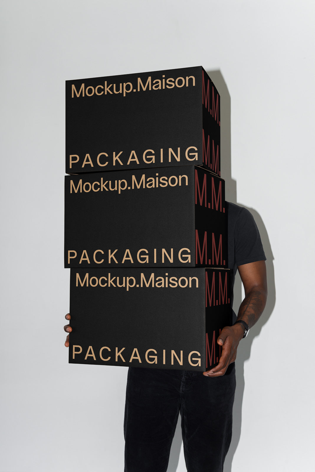PSD mockup of stacked black boxes labeled "Mockup.Maison" and "PACKAGING" held by a person with a neutral background.