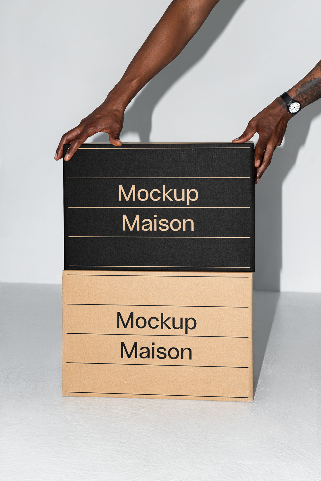PSD mockup of two boxes with "Mockup Maison" text, one black and one cardboard-colored, with hands adjusting the top box against a light background.