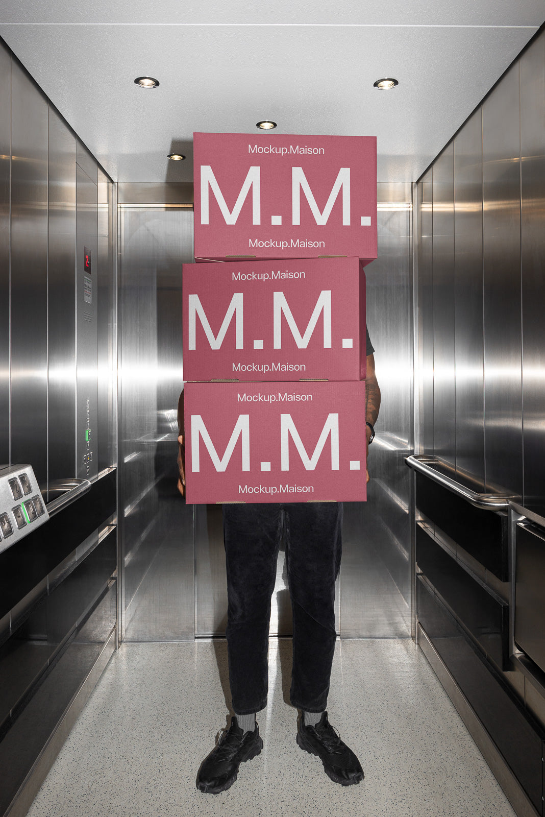 PSD mockup of a person standing in an elevator holding four stacked boxes labeled "Mockup Maison" with large white "M.M." text.