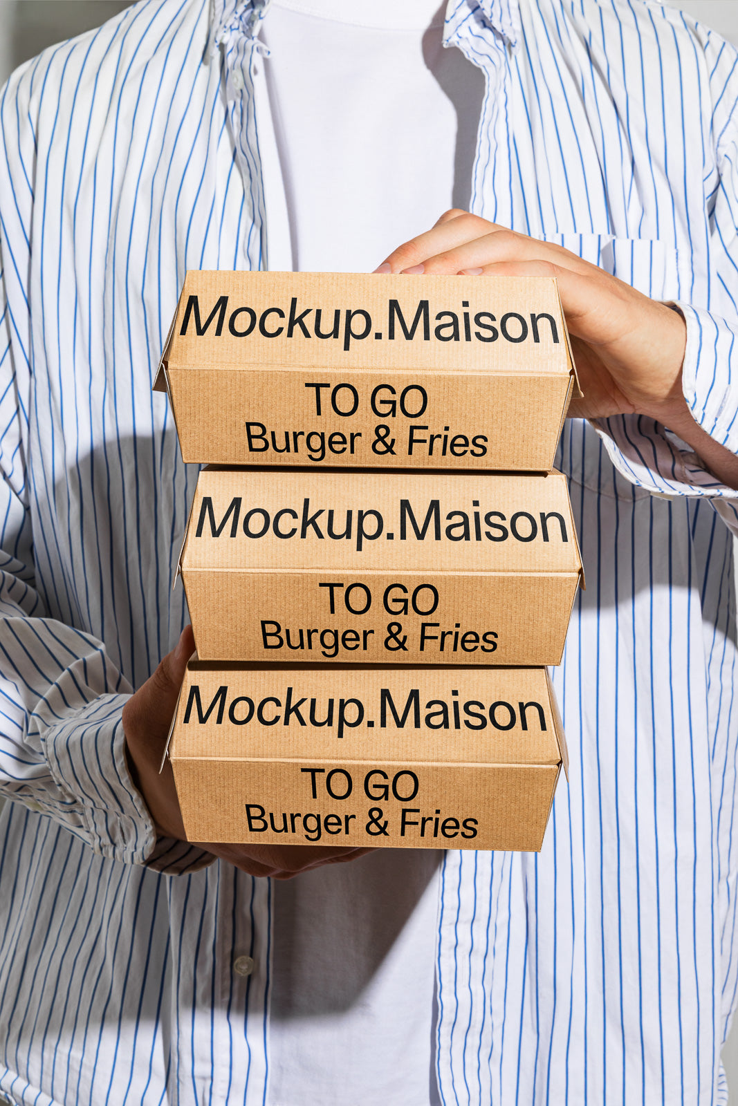 PSD mockup of a person holding three stacked cardboard takeout boxes labeled "Mockup.Maison TO GO Burger & Fries," wearing a striped shirt.