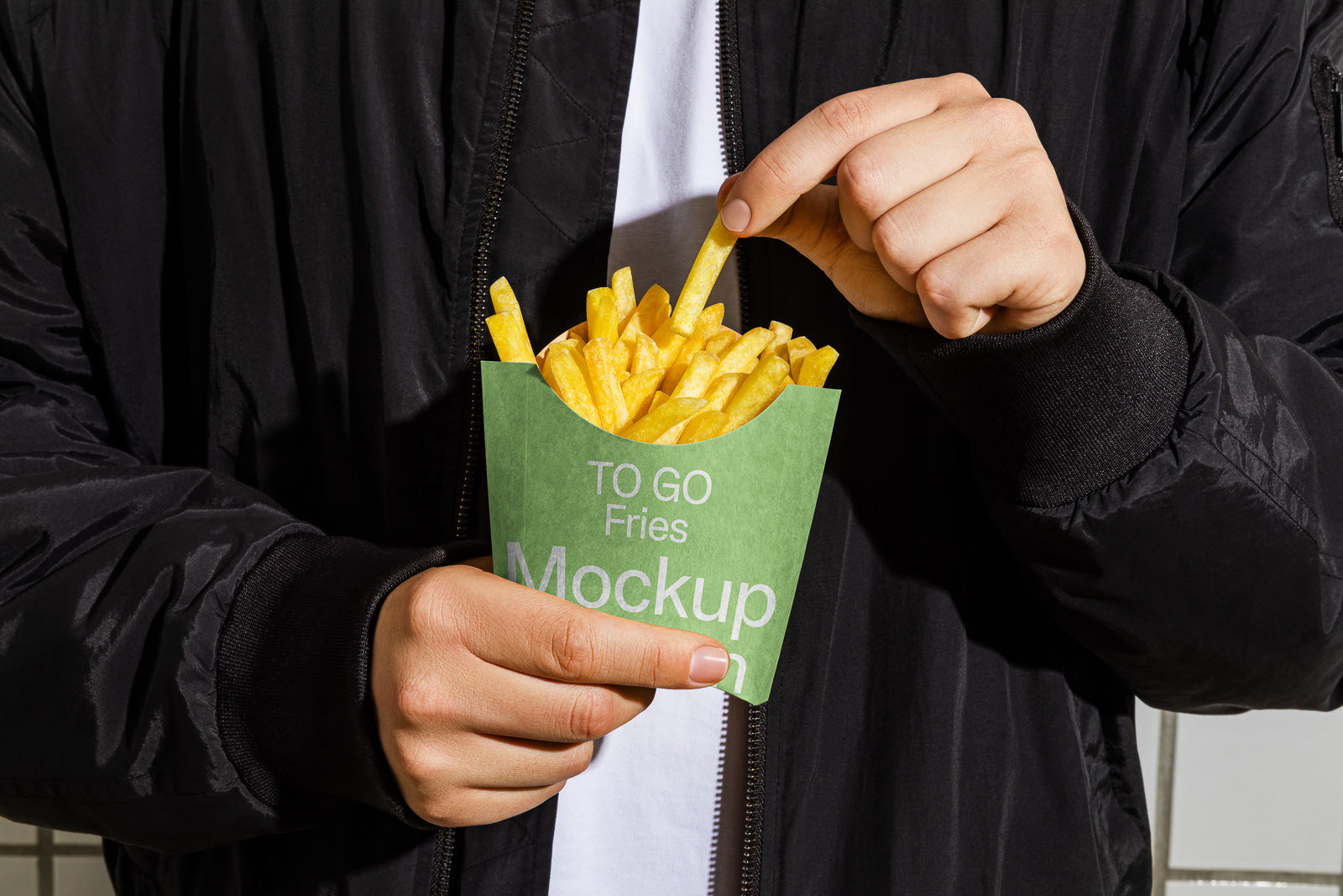 PSD mockup of a person holding a green carton labeled "TO GO Fries" filled with golden fries, wearing a black jacket and white shirt.