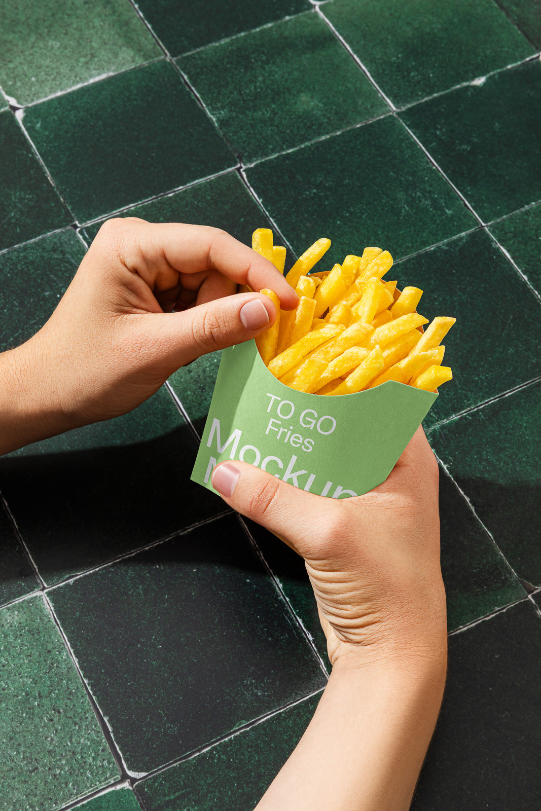 PSD mockup of hands holding a green fries container labeled "To Go Fries" against a dark green tiled surface.