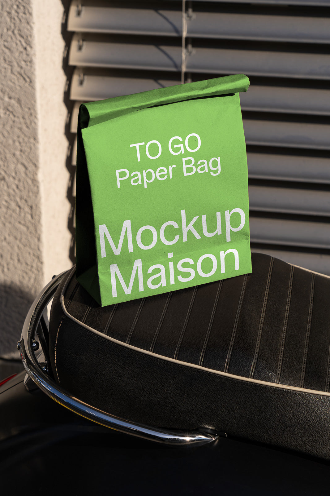 PSD mockup of a green to-go paper bag labeled "Mockup Maison" positioned on a black leather seat with striped stitching, set against a gray shutter background.