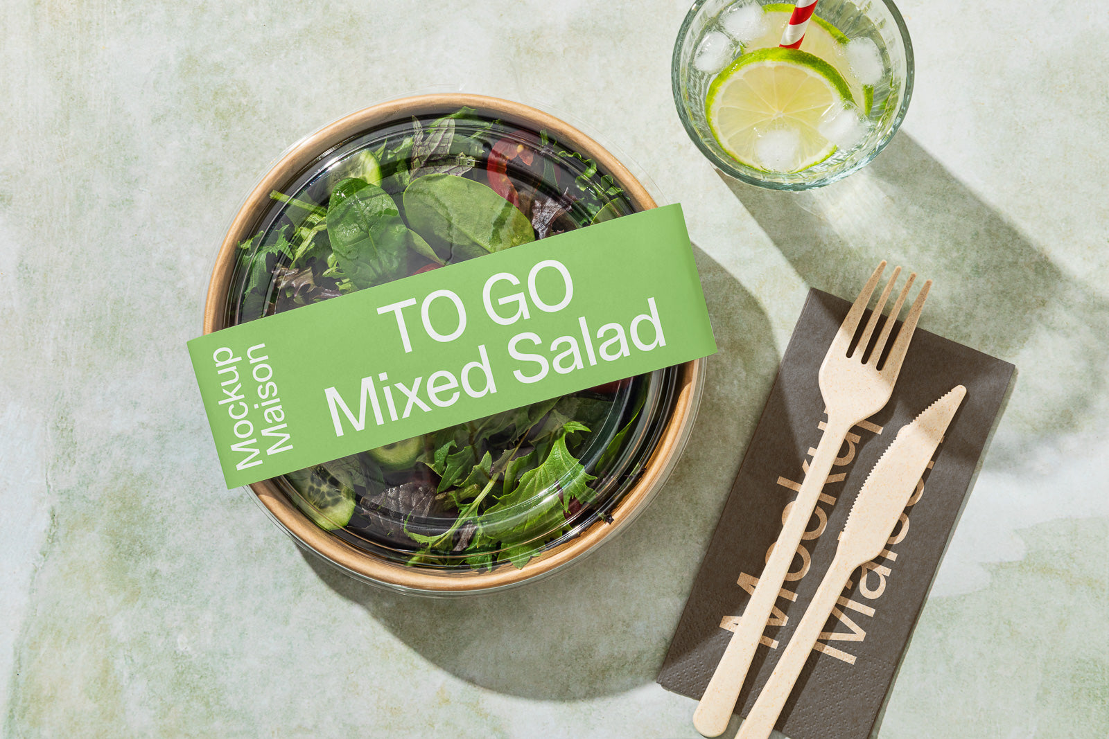 PSD mockup of a takeaway salad in a round container with a clear lid, labeled "TO GO Mixed Salad," alongside a glass of lime water and wooden cutlery on a napkin.
