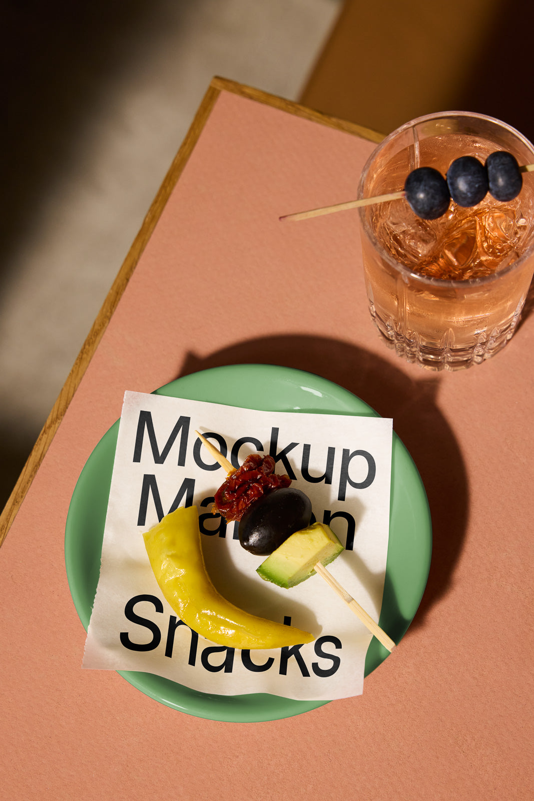 PSD mockup of a green plate with a fruit skewer and decorative napkin, accompanied by a glass of iced pink drink with blueberries on a skewer.