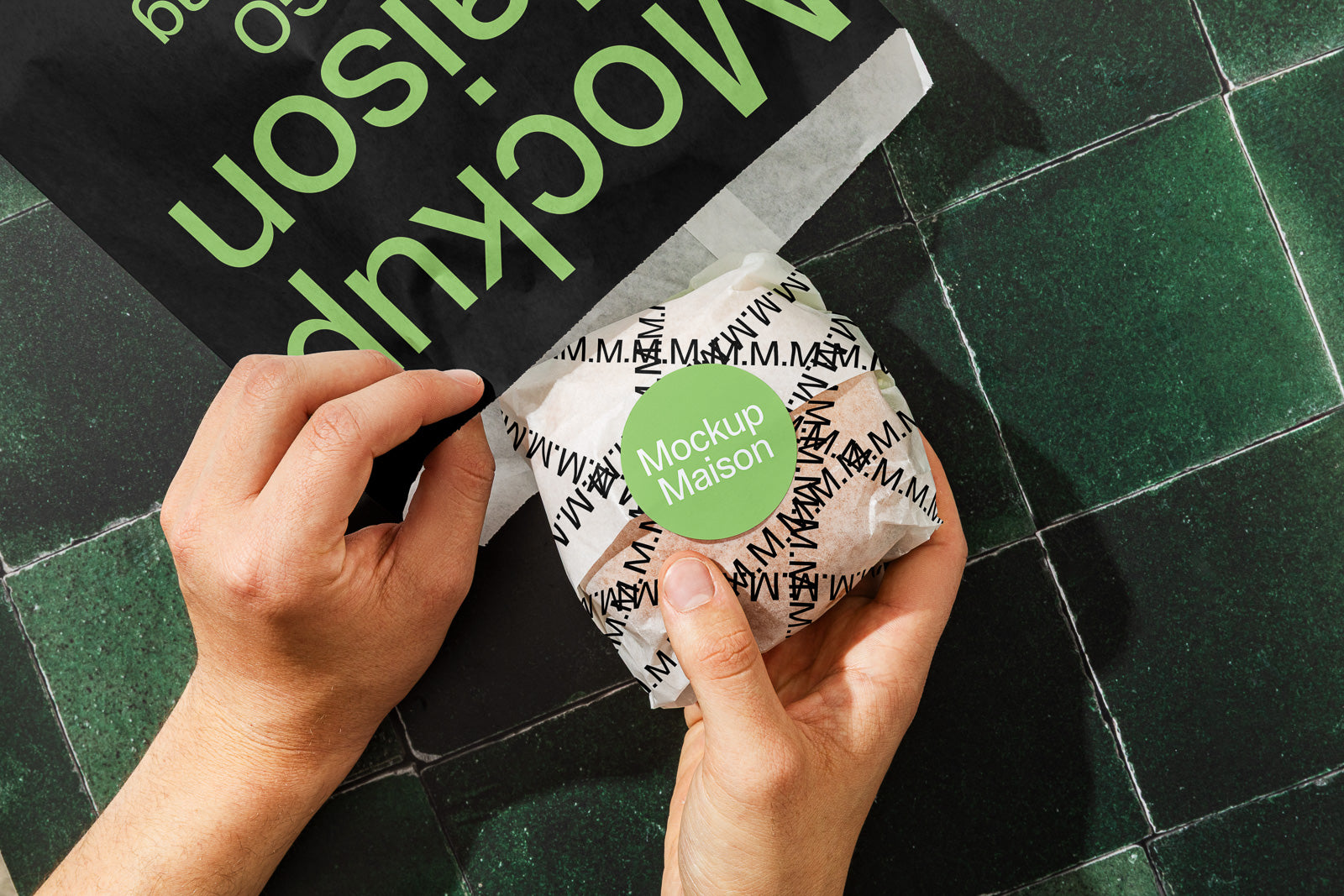PSD mockup of hands holding a branded sandwich wrap with "Mockup Maison" sticker on black and white pattern, next to a black paper bag with green text on a tiled surface.