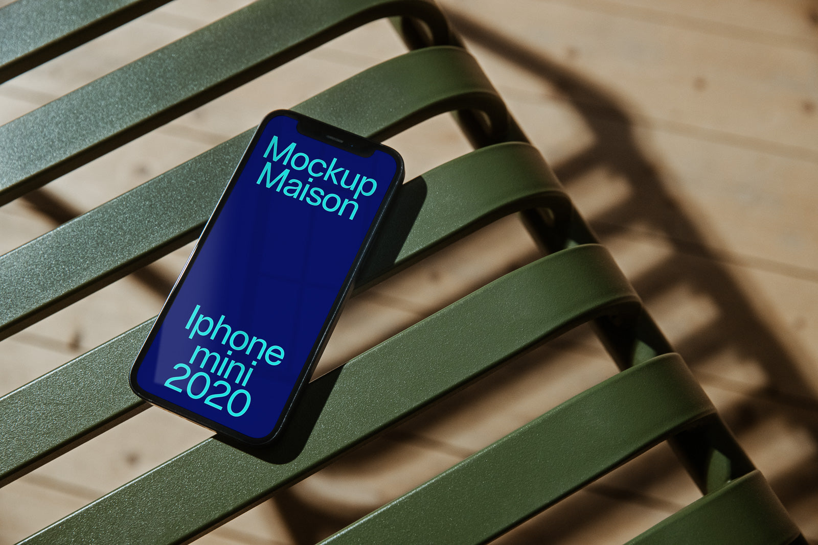 PSD mockup of an iPhone Mini 2020 lying on a green metal chair with sunlight casting shadows, displaying "Mockup Maison" on its screen.