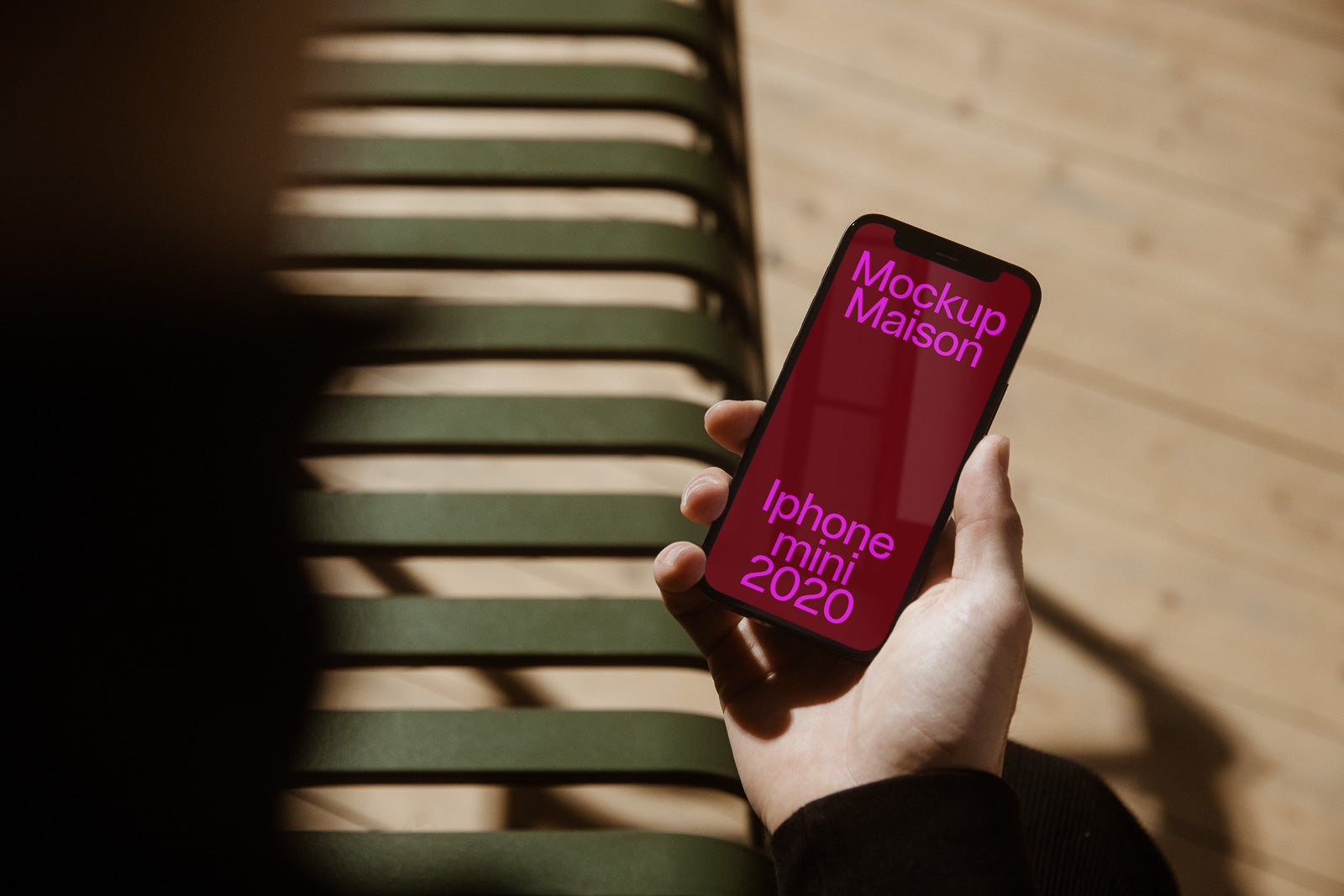 PSD mockup of a person holding an iPhone Mini 2020 with "Mockup Maison" text on screen, sitting on a slatted bench with shadows.